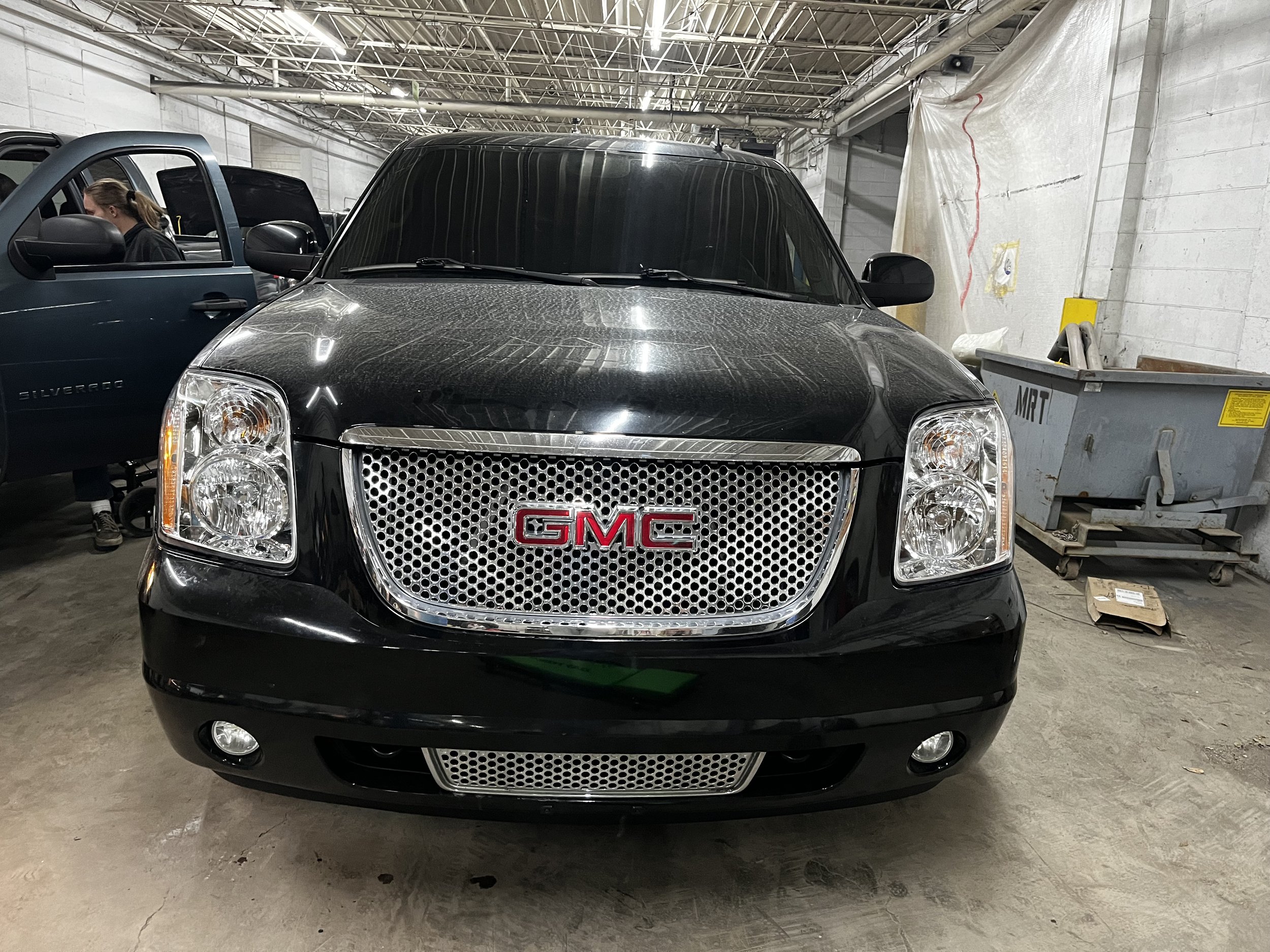 Front view of a black GMC SUV parked in a garage with a nearby blue SUV and a dumpster in the background.