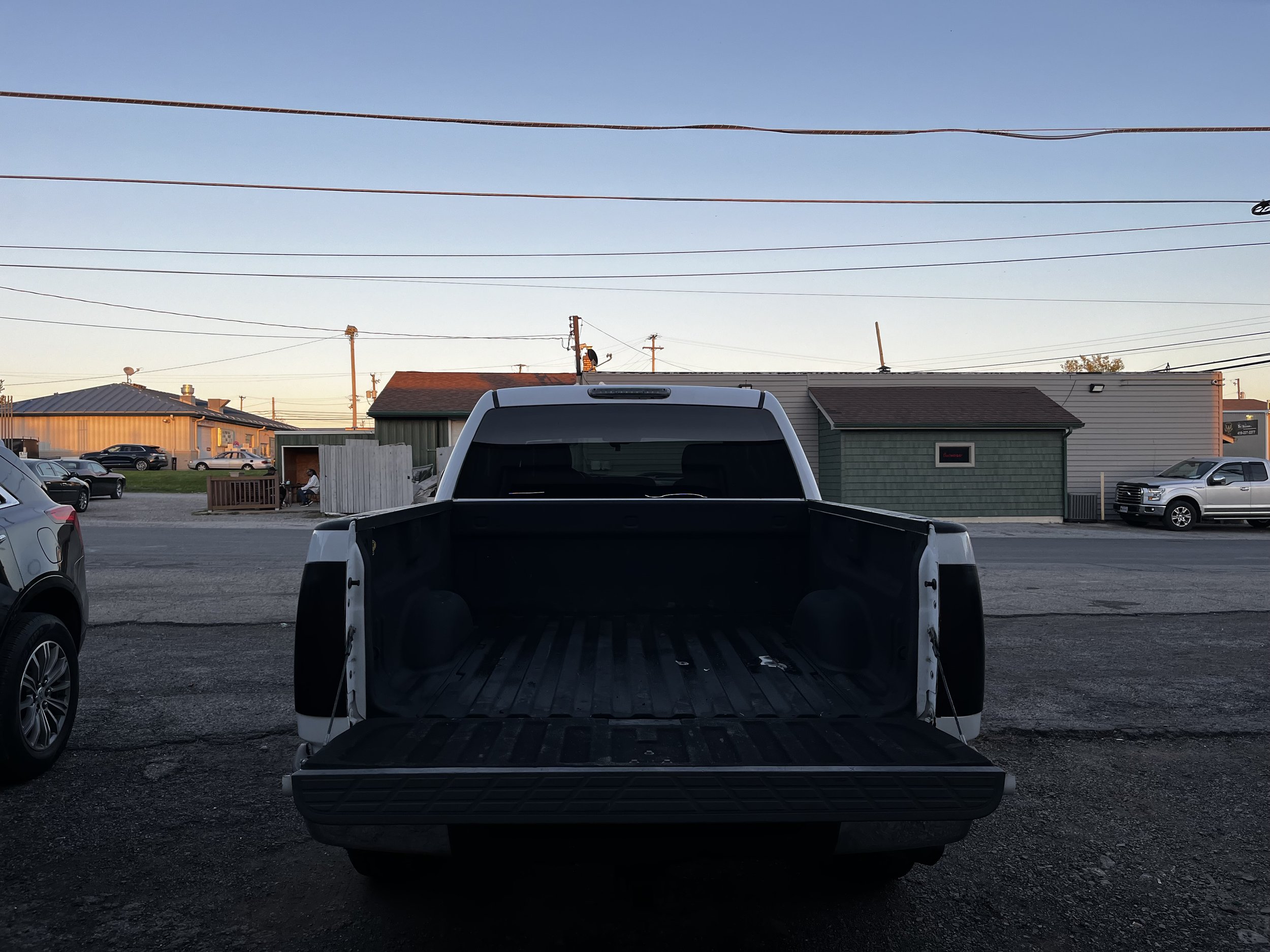 Open bed of a pickup truck parked in a lot with buildings and parked cars in the background.