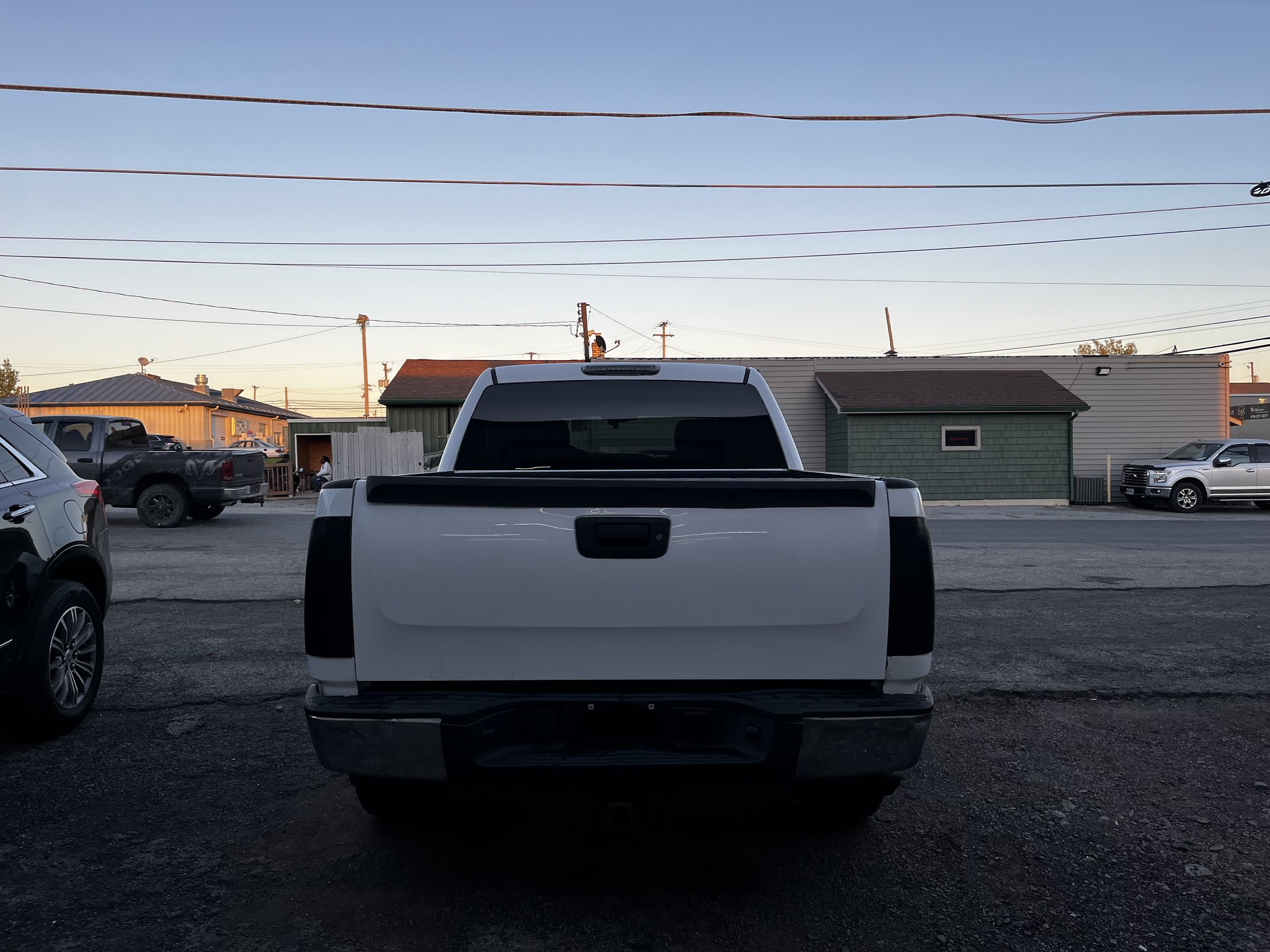 Rear view of a white pickup truck in a parking lot, surrounded by other vehicles and buildings in the background.