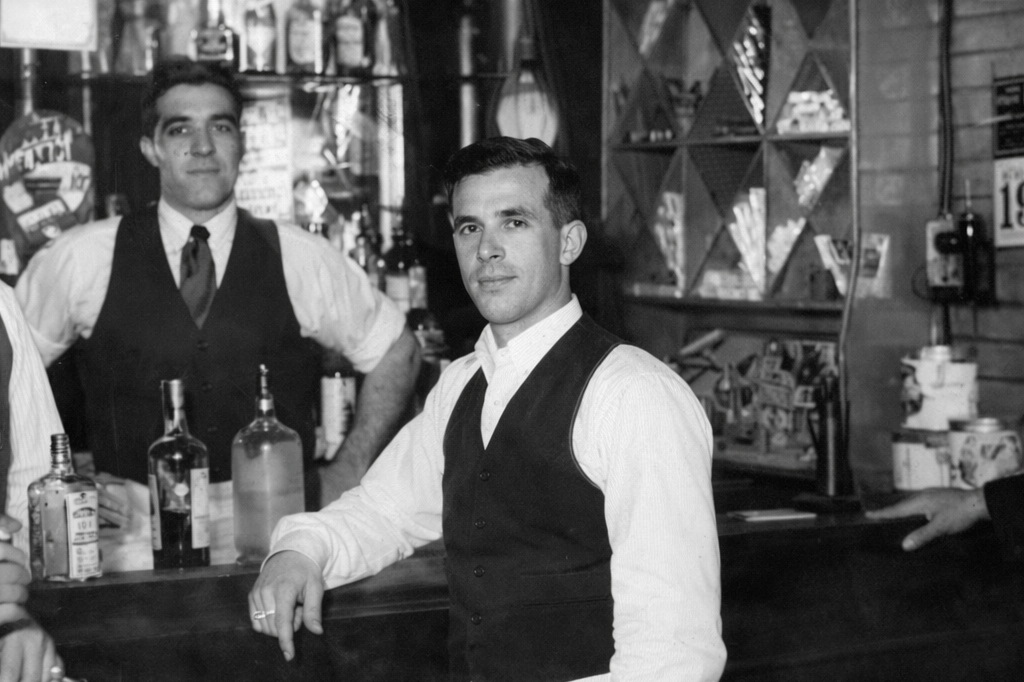 Two bartenders behind a bar, with liquor bottles and bar supplies, in a vintage black-and-white setting.