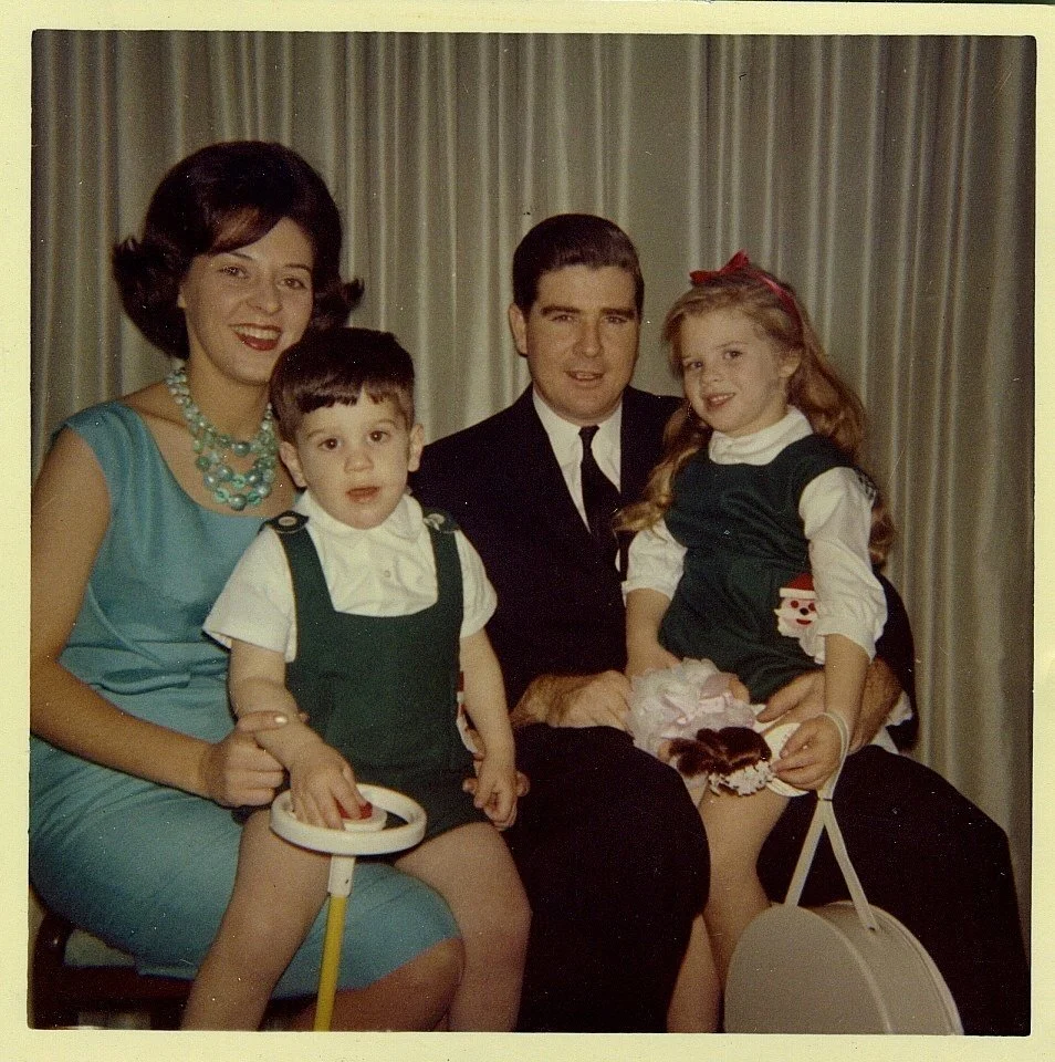 A retro family portrait showing a woman with dark hair in a blue dress and pearl necklace, a man in a black suit, a young boy in a white shirt and green overalls, and a girl in a Christmas-themed dress with a red bow, sitting in front of beige curtains.