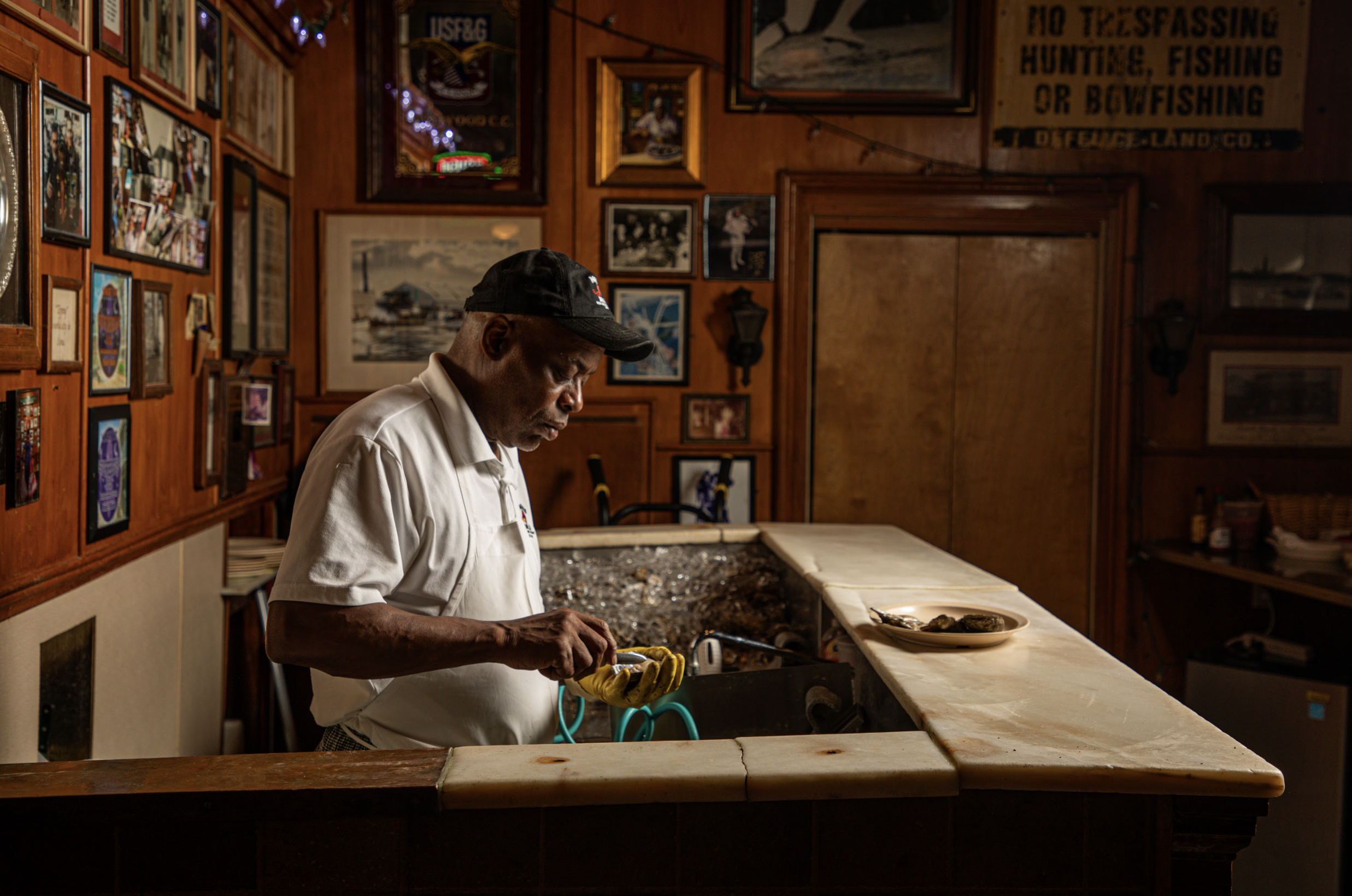 An elderly man wearing a black cap and white shirt stands behind a counter in a wooden-furnished restaurant or bar, preparing food or a drink. The wall behind him is decorated with various framed photos, painting, and memorabilia, giving a cozy, nostalgic atmosphere.