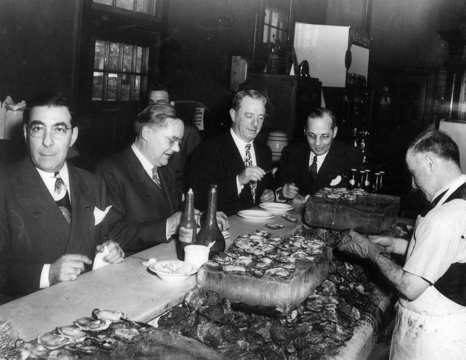 Men in suits enjoying oysters at a seafood bar, with a worker shucking oysters on ice.