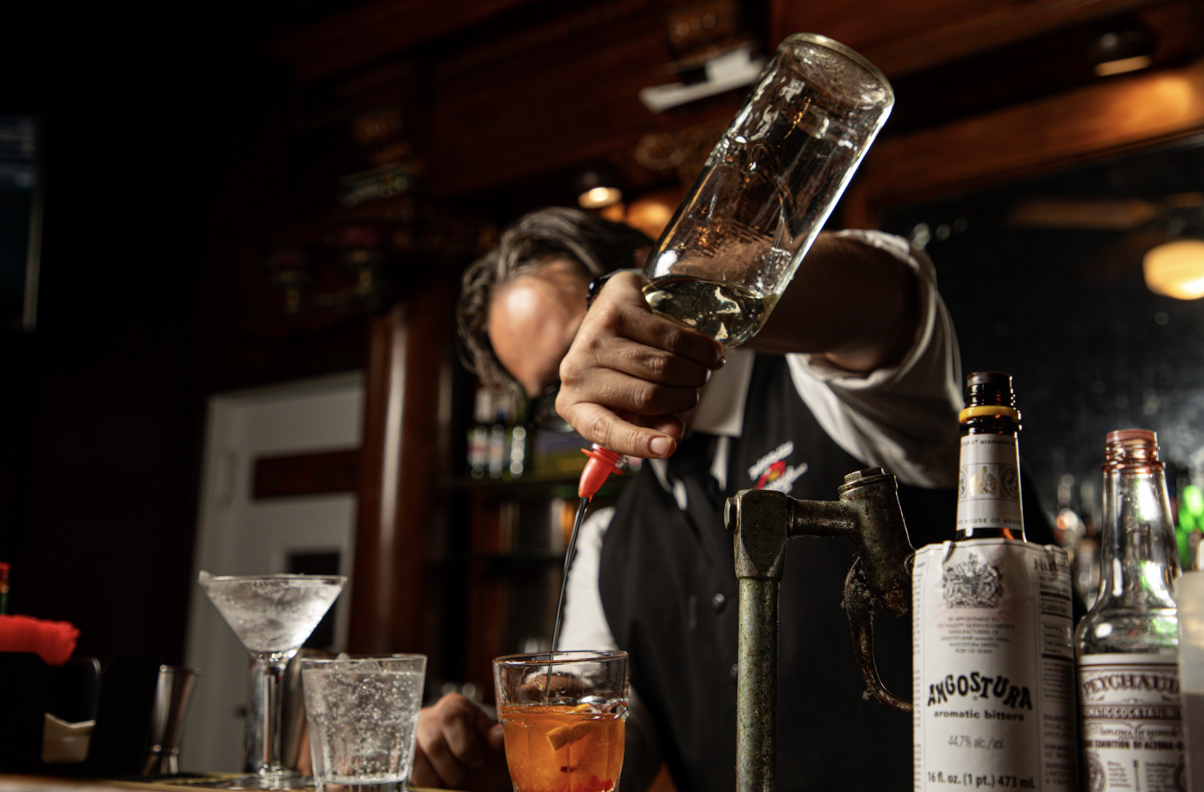 Bartender pouring a drink into a glass at a bar with various bottles, glasses, and bar tools on the counter.