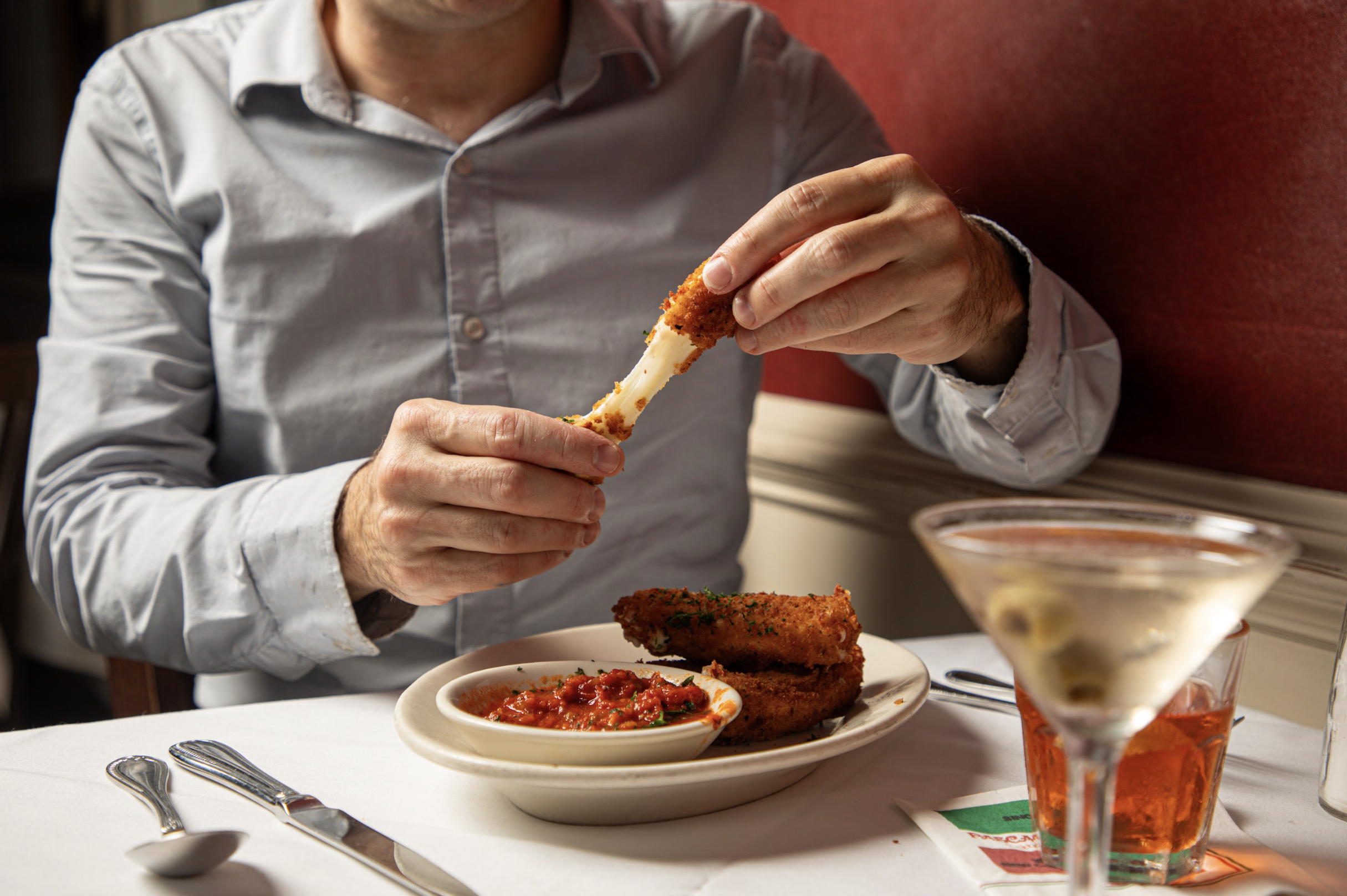 A man in a white shirt is eating a piece of fried chicken with cheese inside. There are bowls of tomato-based sauce and other dishes on the table, along with a glass of an orange-colored beverage.