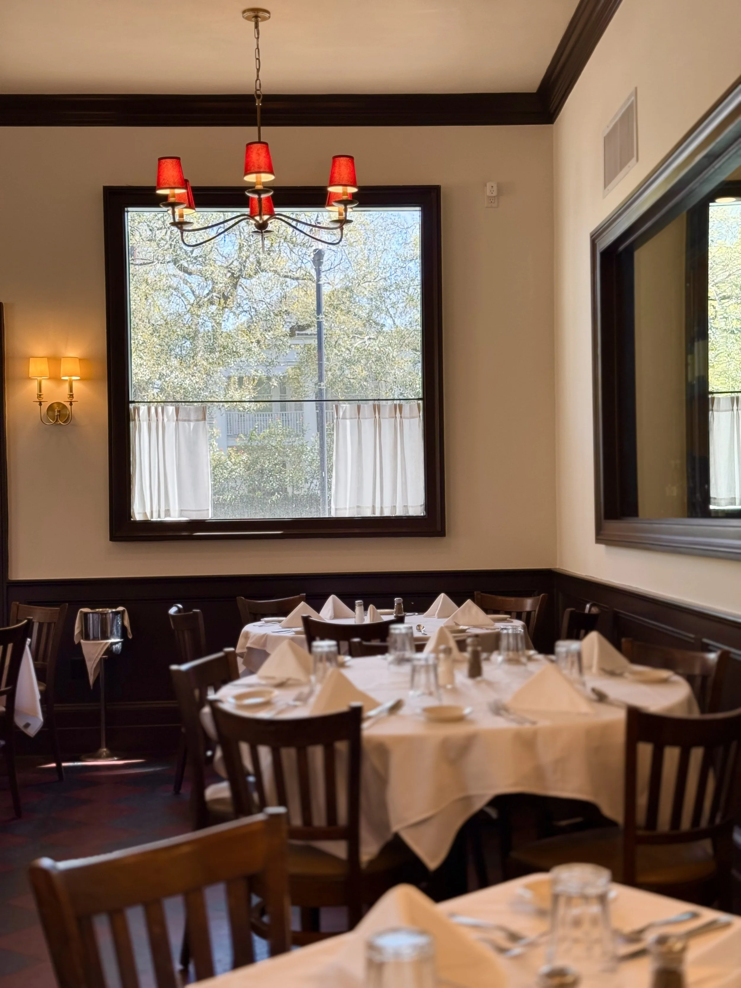 Interior of a restaurant with multiple round tables covered with white tablecloths, set with napkins, glasses, and cutlery, surrounded by wooden chairs. A large window with curtains overlooks trees outside. Red chandelier hanging from the ceiling, and wall-mounted lights with yellow shades.