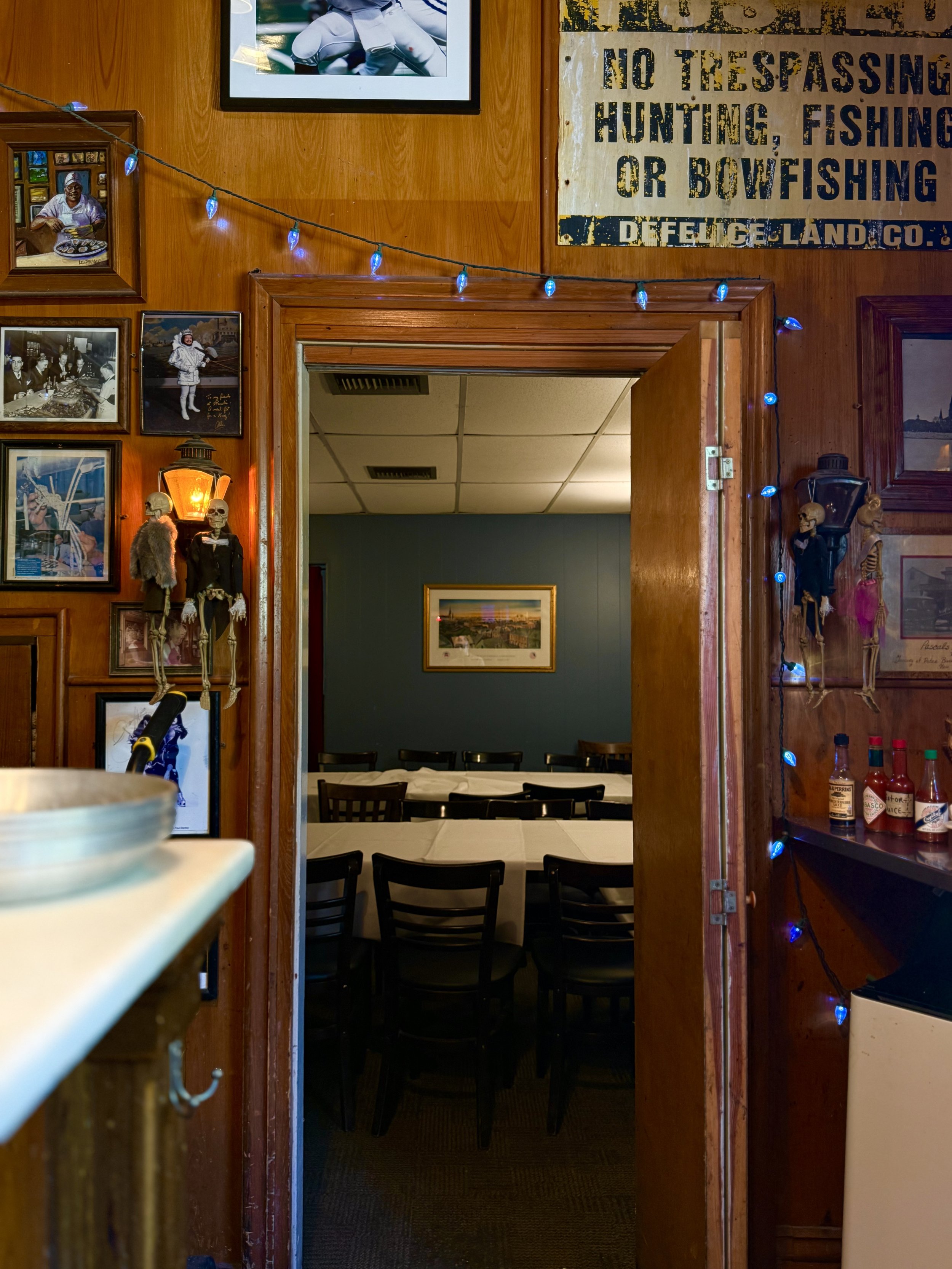 A view through a restaurant doorway showing tables and chairs inside, wall art, and framed pictures, with some skeleton decorations and blue string lights on the wooden walls.