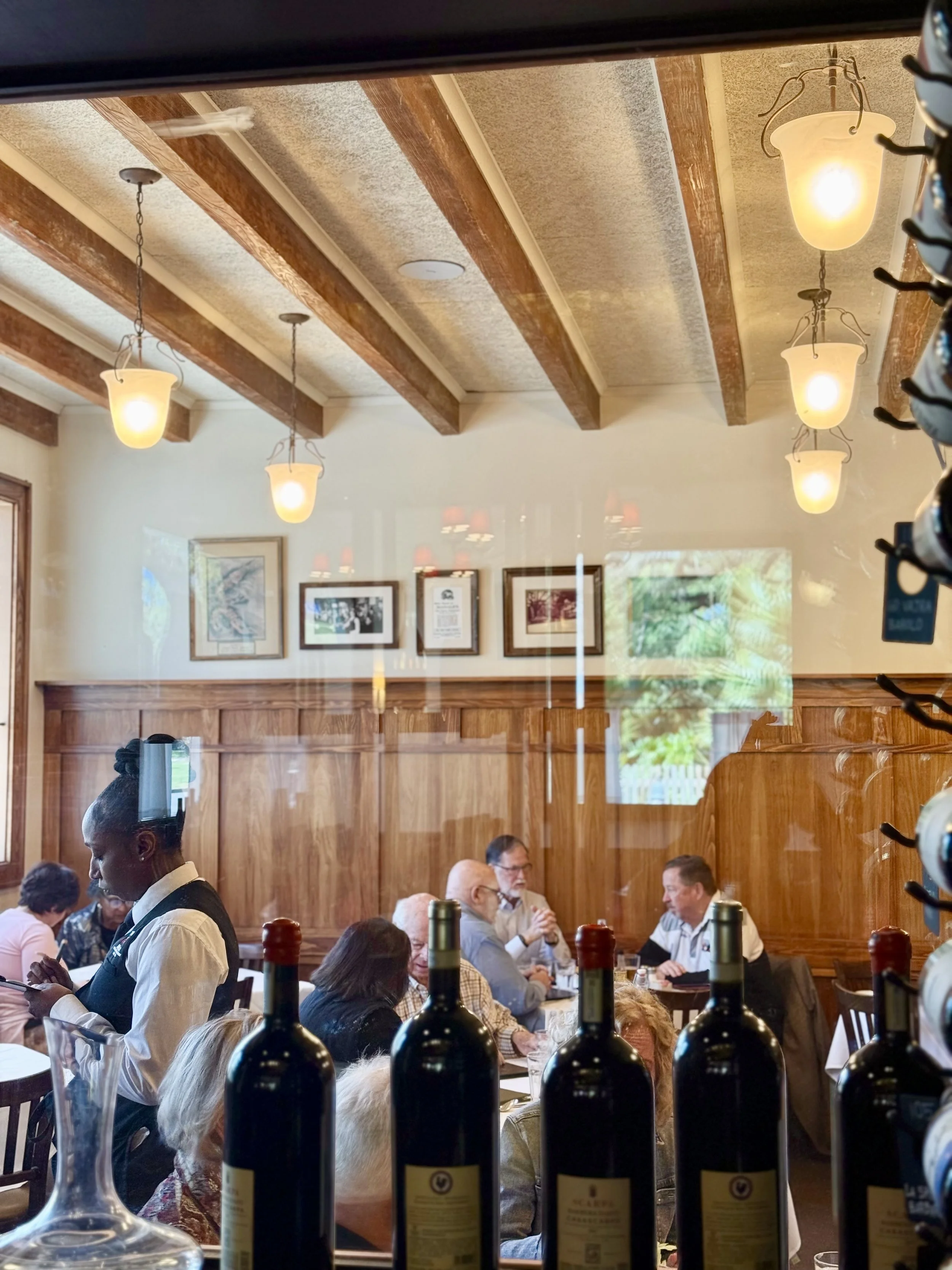 A busy restaurant with people dining and a waitress serving food. The scene is viewed through a window or mirror, with wine bottles and glasses in the foreground. The ceiling features wooden beams and hanging lights.