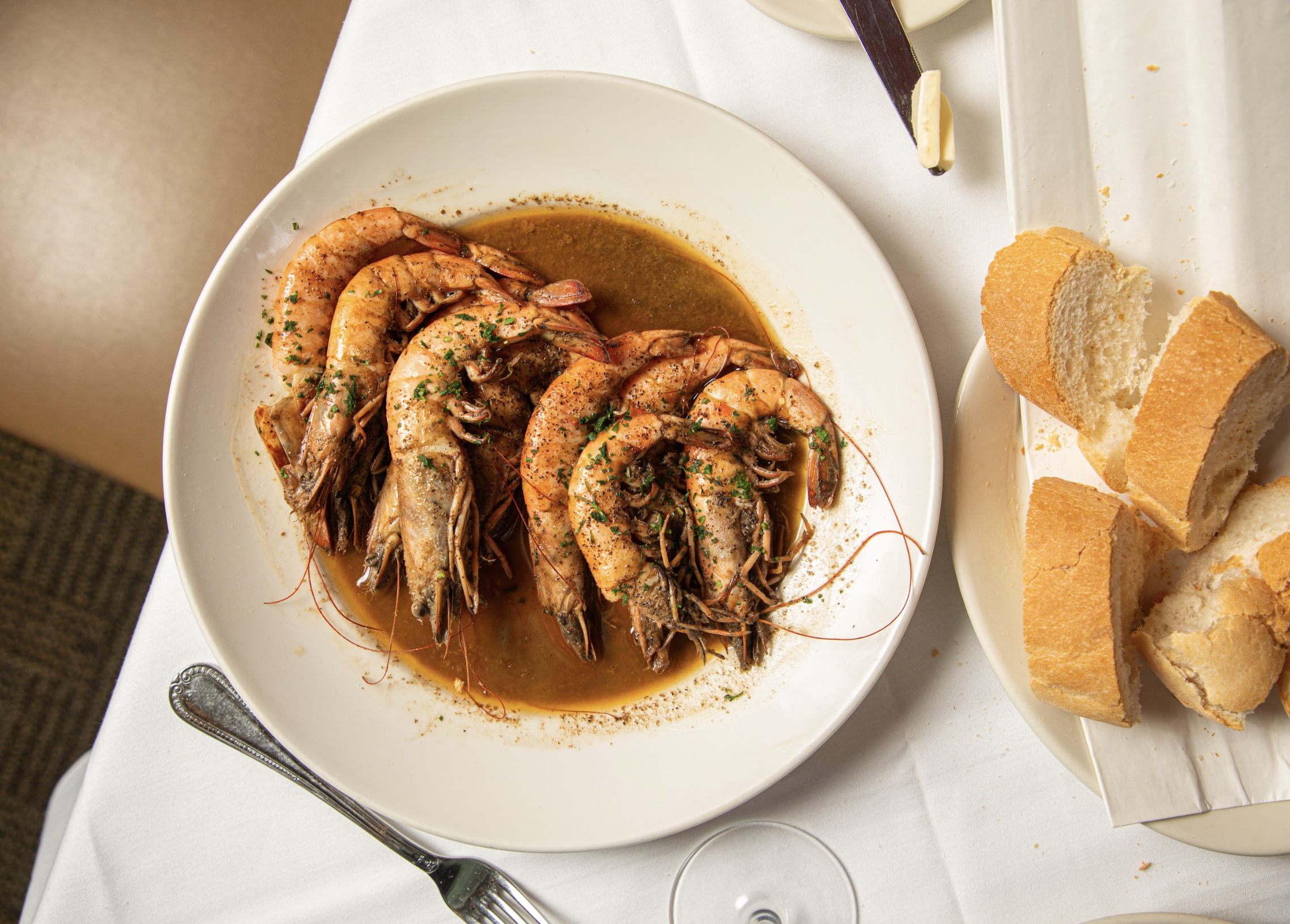 A white plate contains cooked shrimp garnished with herbs and surrounded by a sauce. In the background, there are slices of bread on a separate white dish, and a glass of white wine is partially visible.