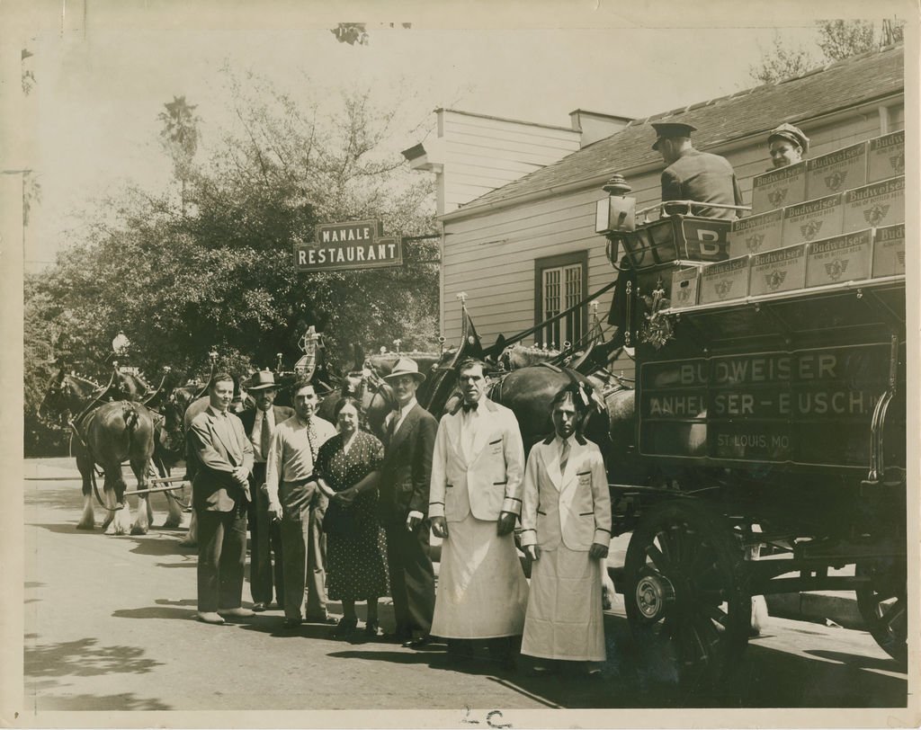 Black and white vintage photo of people standing on a street in front of a horse-drawn wagon loaded with beer cases, with a building and trees in the background.