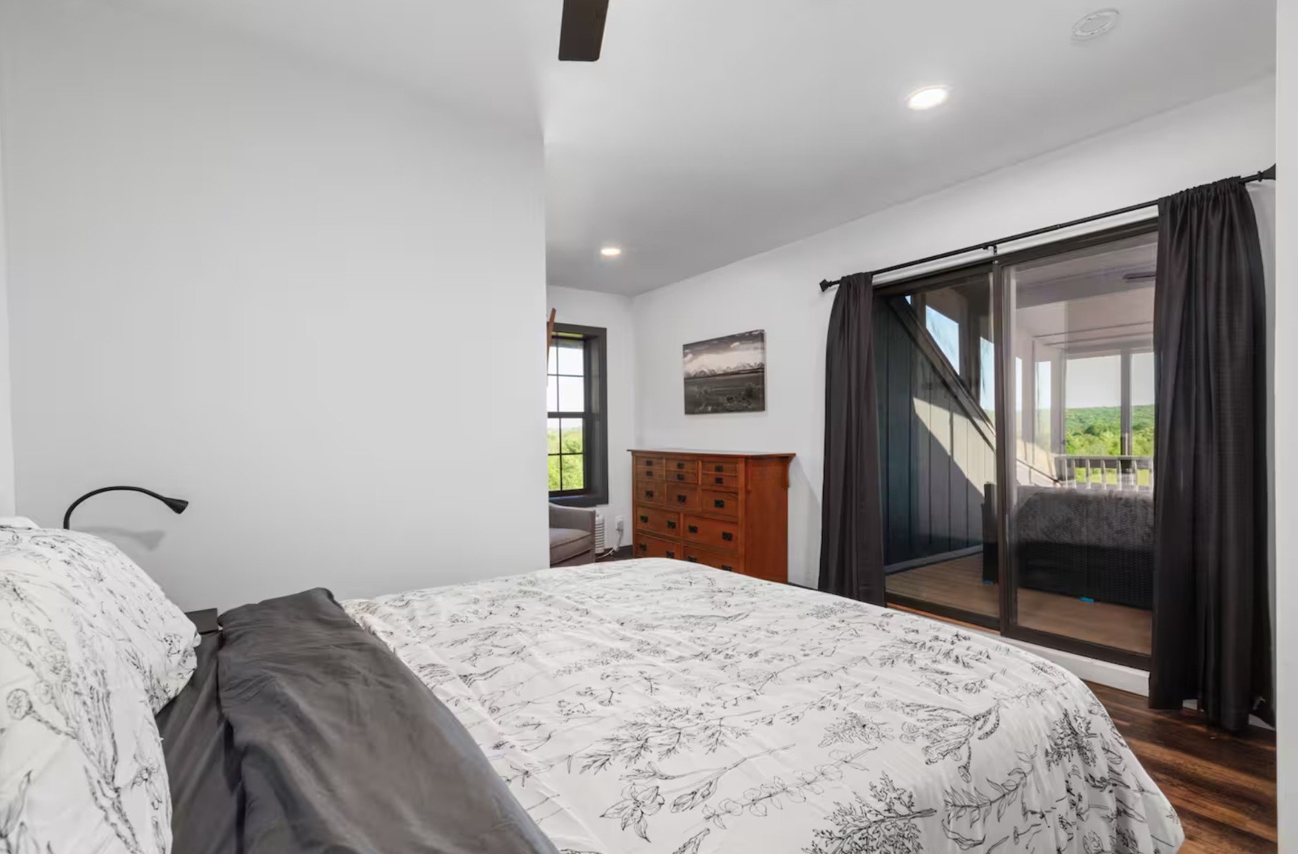 Bedroom with white walls, large bed with white floral bedding, dark gray pillow, black reading lamp, wooden dresser, window with black trim, and sliding glass door with dark curtains leading to a balcony with green landscape outside.