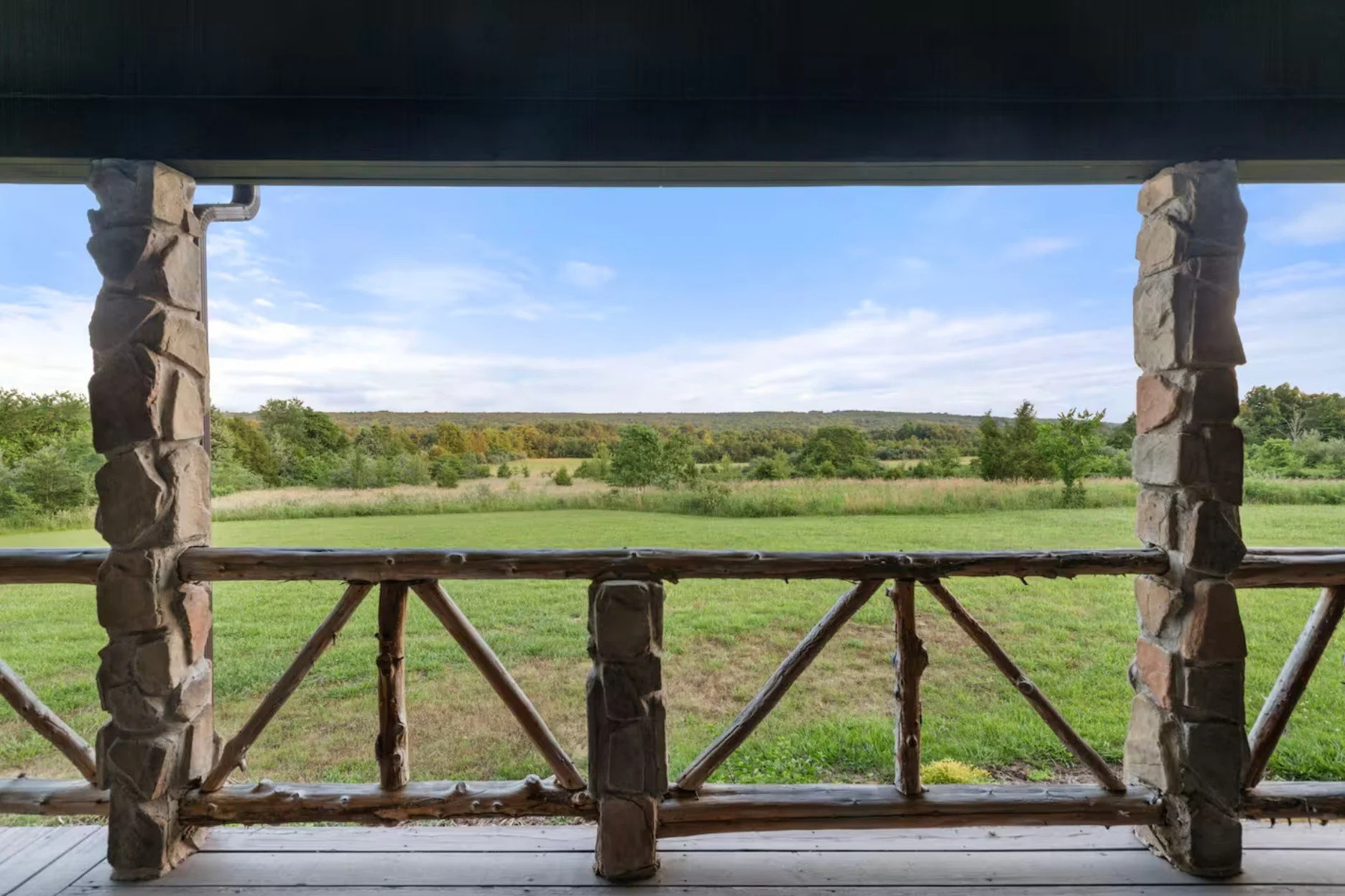 Rural landscape viewed from a porch with stone pillars and wooden railing; green fields, trees, and a blue sky with clouds.
