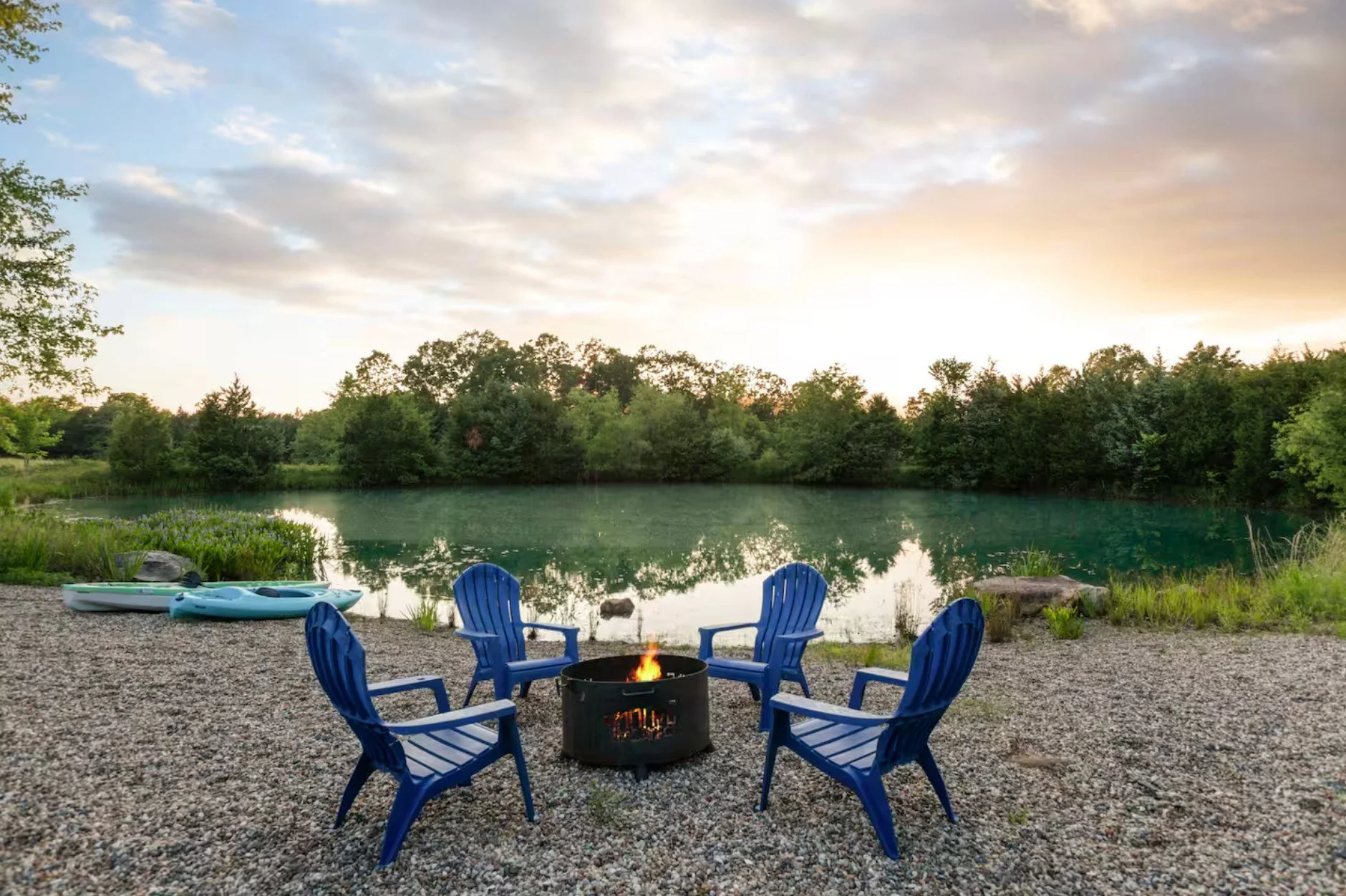 Four blue Adirondack chairs arranged in a circle around a fire pit on a pebble beach near a lake, with two kayaks on the shore and lush green trees in the background.