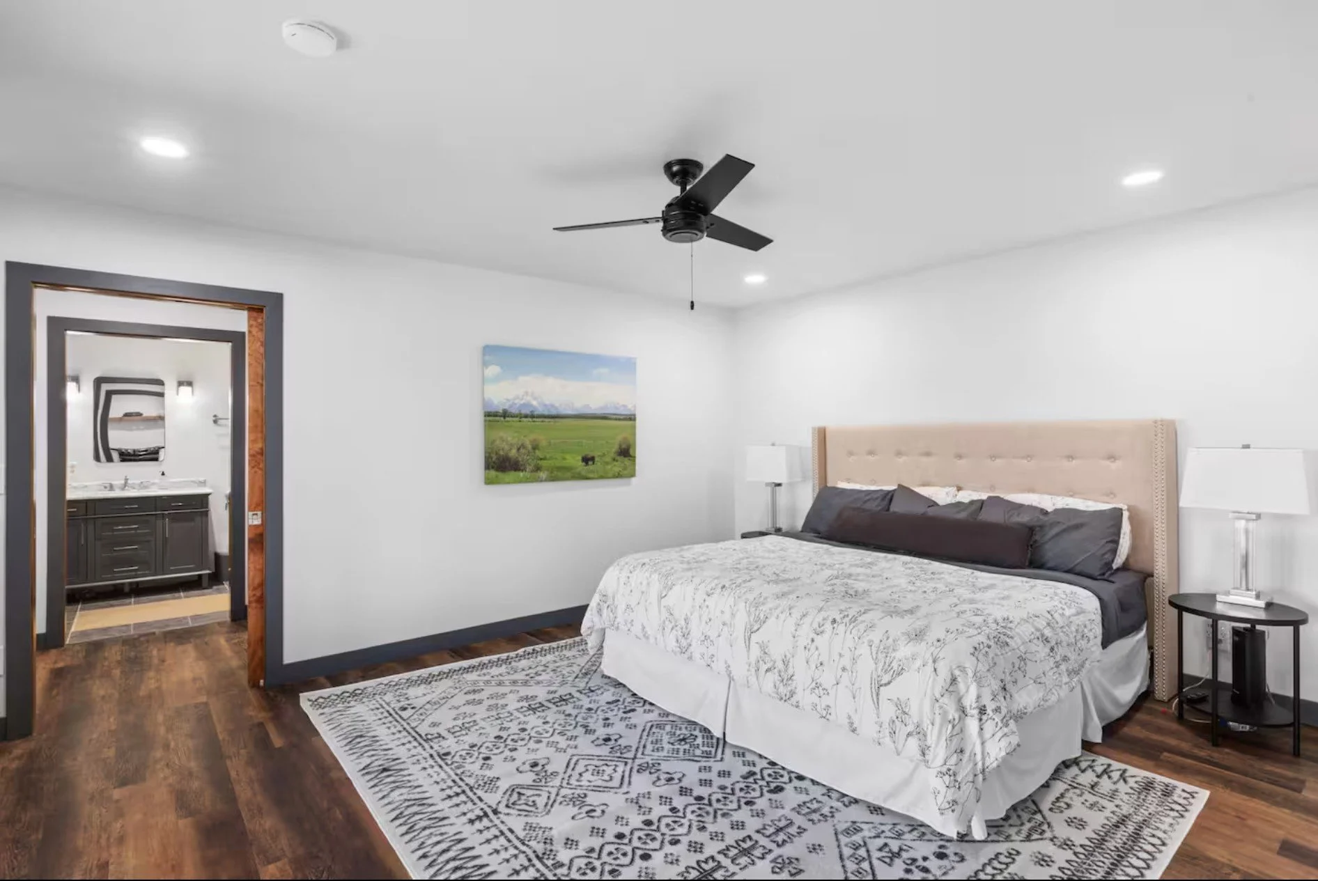 A bedroom with a large bed, white bedding, and black pillows, with a side table and lamp, a ceiling fan, wood flooring, and a corridor leading to a bathroom with a dark vanity and mirror.
