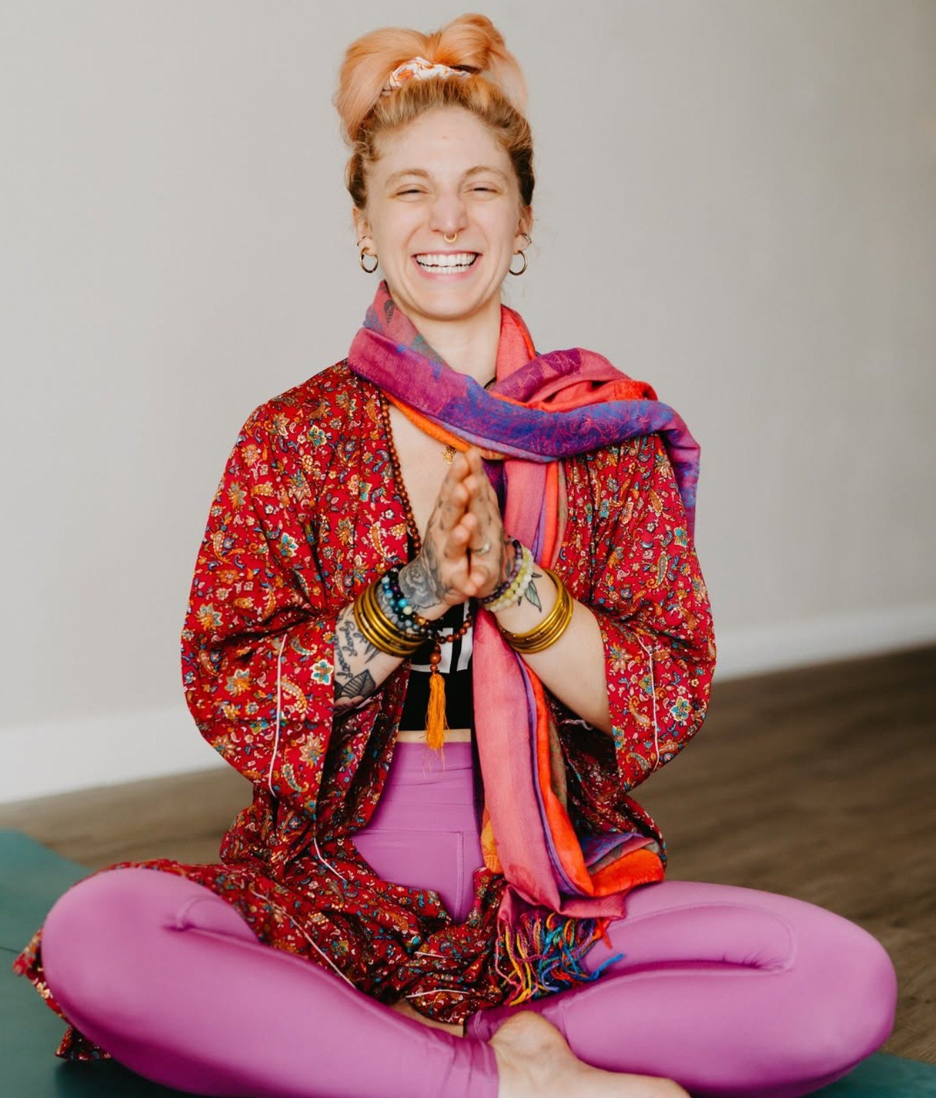 A woman with pink hair tied up, wearing a colorful scarf, red patterned top, pink leggings, and bracelets, sitting cross-legged on the floor, smiling and praying with clasped hands.