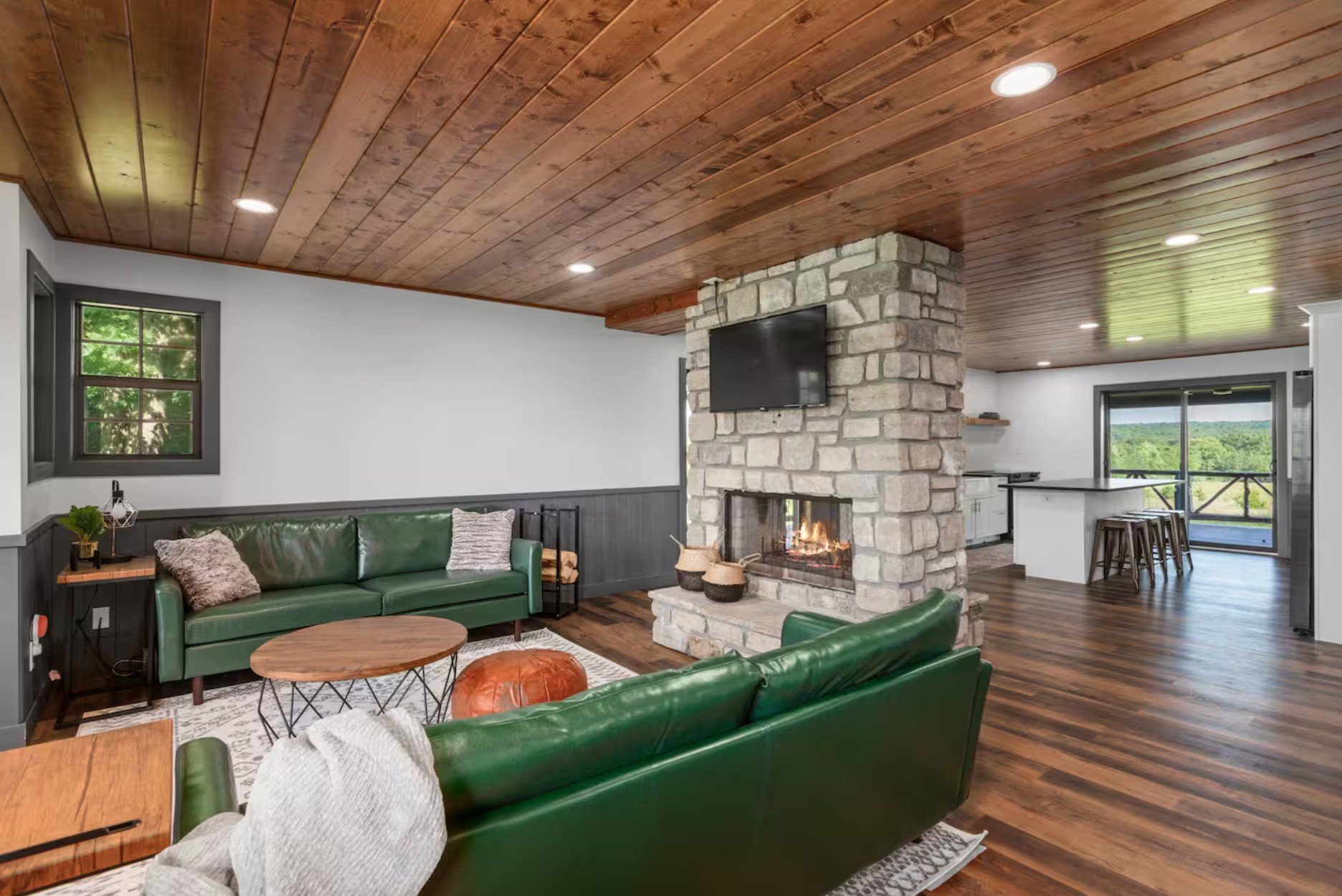Living room with green leather sofas, wooden coffee table, stone fireplace with a mounted TV above, wood-panel ceiling, hardwood floor, and sliding glass door leading to an outdoor view.