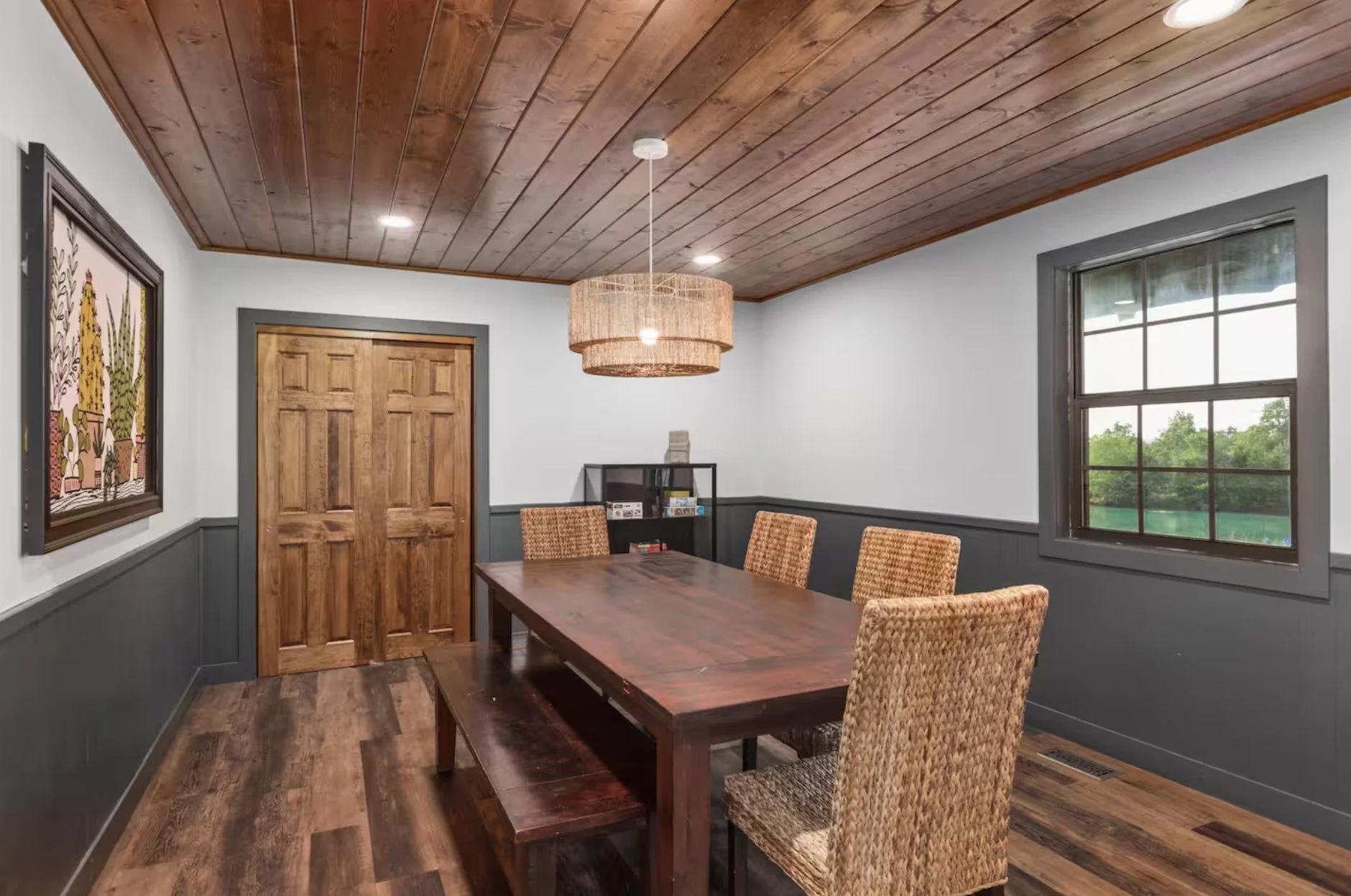 A dining room with a wooden table and four woven chairs, wood-paneled ceiling, gray walls with dark gray trim, a window showing green trees outside, framed artwork on the wall, a hanging light fixture, and a black bookshelf in the corner.