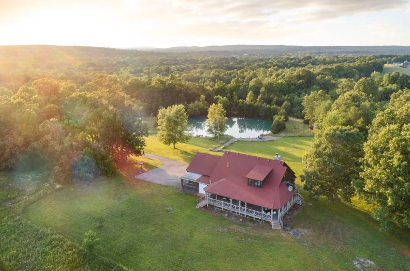 Aerial view of a house with red roof, surrounded by green trees and grass, near a pond, with a driveway and a forest in the background, during sunset.