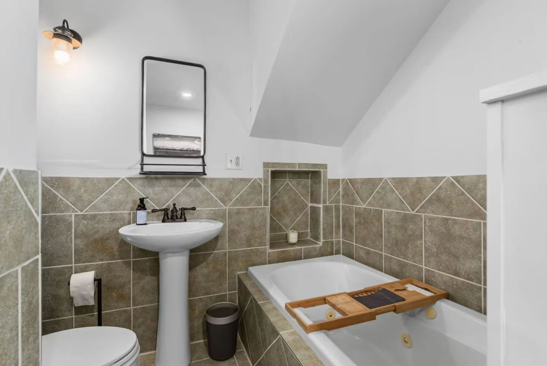 Bathroom with pedestal sink, mirror, toilet, bathtub with wooden tray, beige tiled walls, wall-mounted light, and a small built-in shelf with a candle.