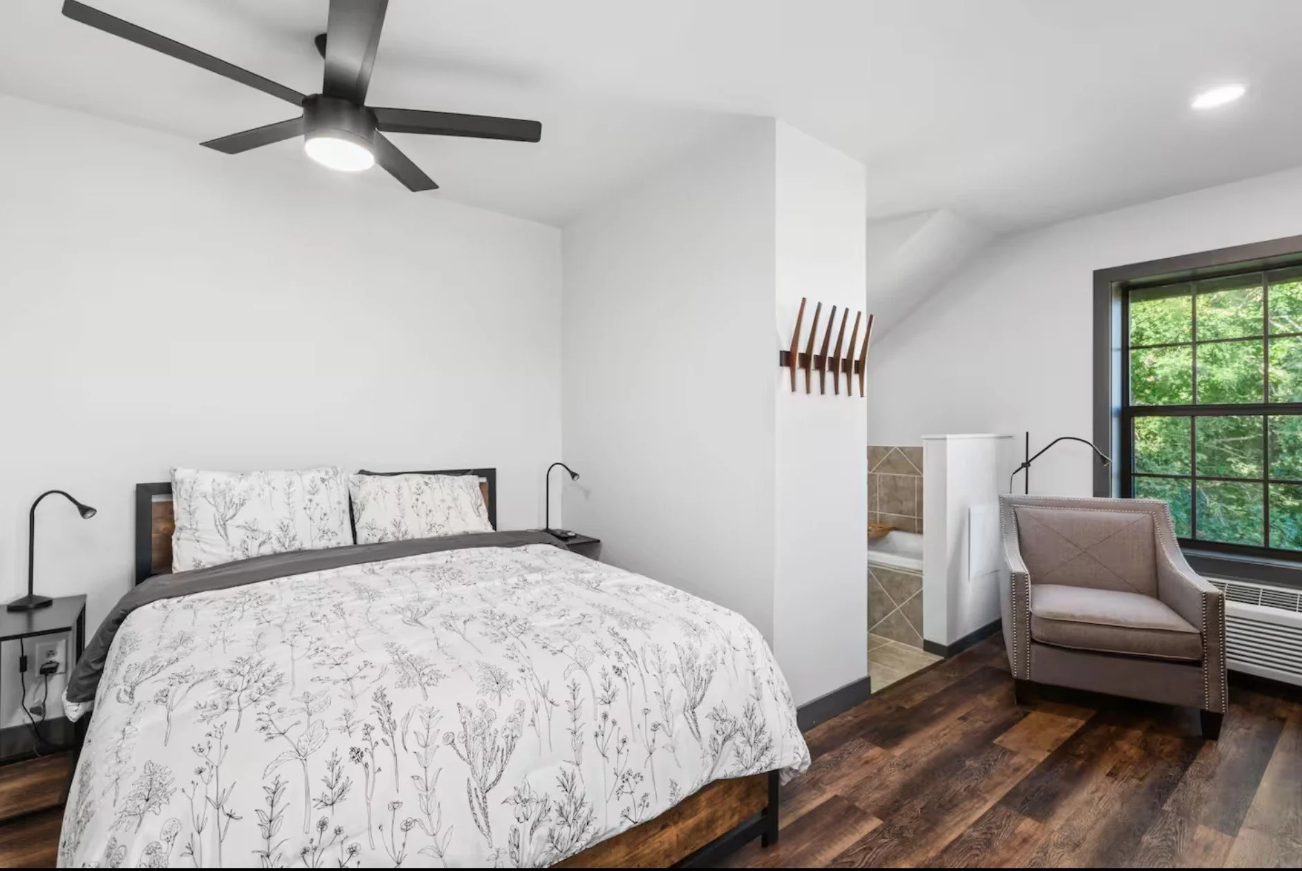 A bedroom with a bed, nightstands, a ceiling fan, an armchair, a window with green trees outside, and a small bathroom or bathtub partially visible.