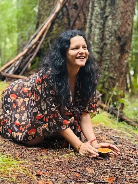 A woman with long, dark, curly hair is outdoors in a forest, sitting on the ground and smiling. She is wearing a dark dress with a colorful leaf and mushroom pattern. She is holding a small, orange mushroom in her hand.