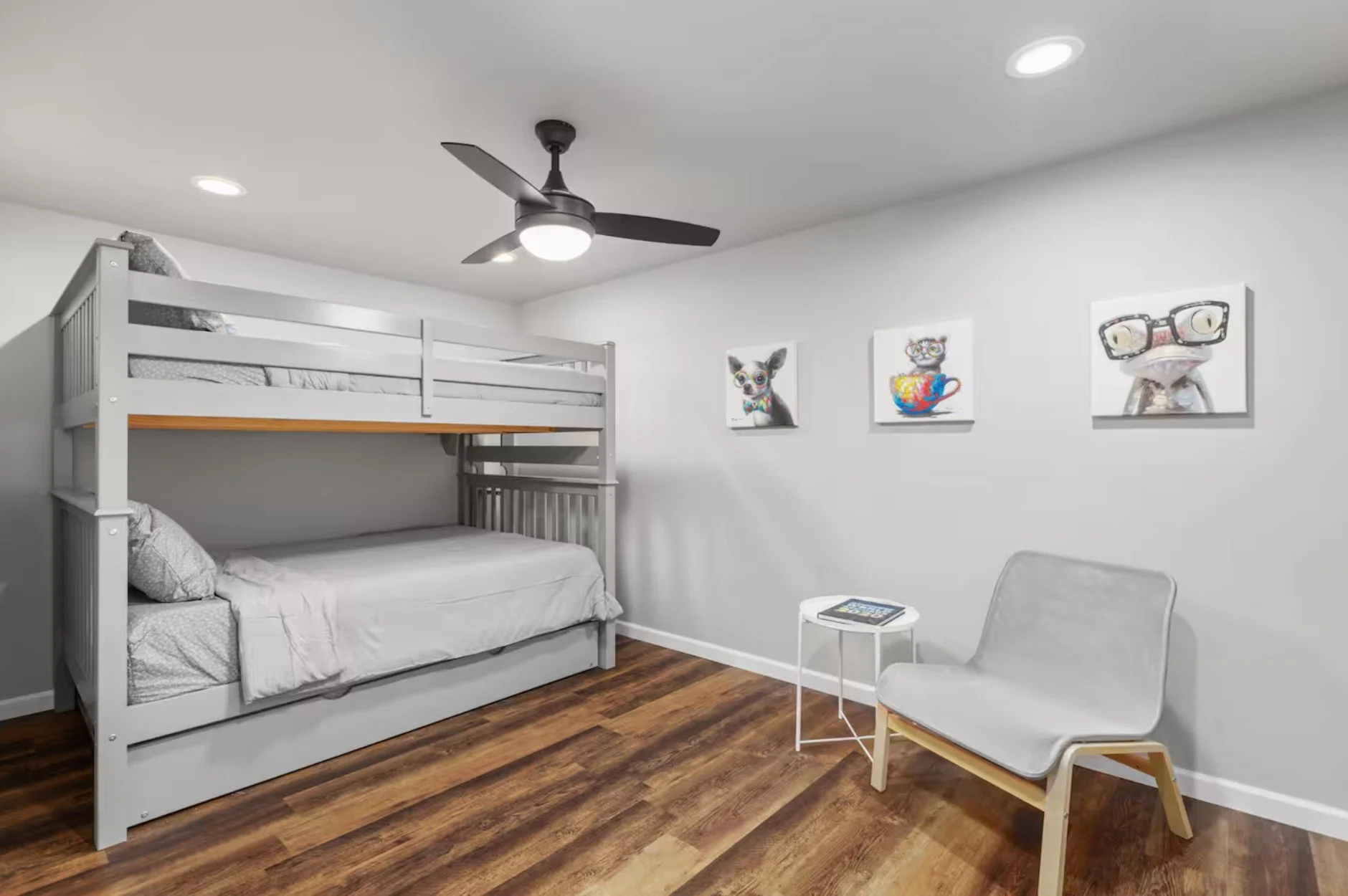 A bedroom with white walls, wooden floor, a white bunk bed, a white chair, a small white side table with a book, and three colorful animal paintings on the wall.