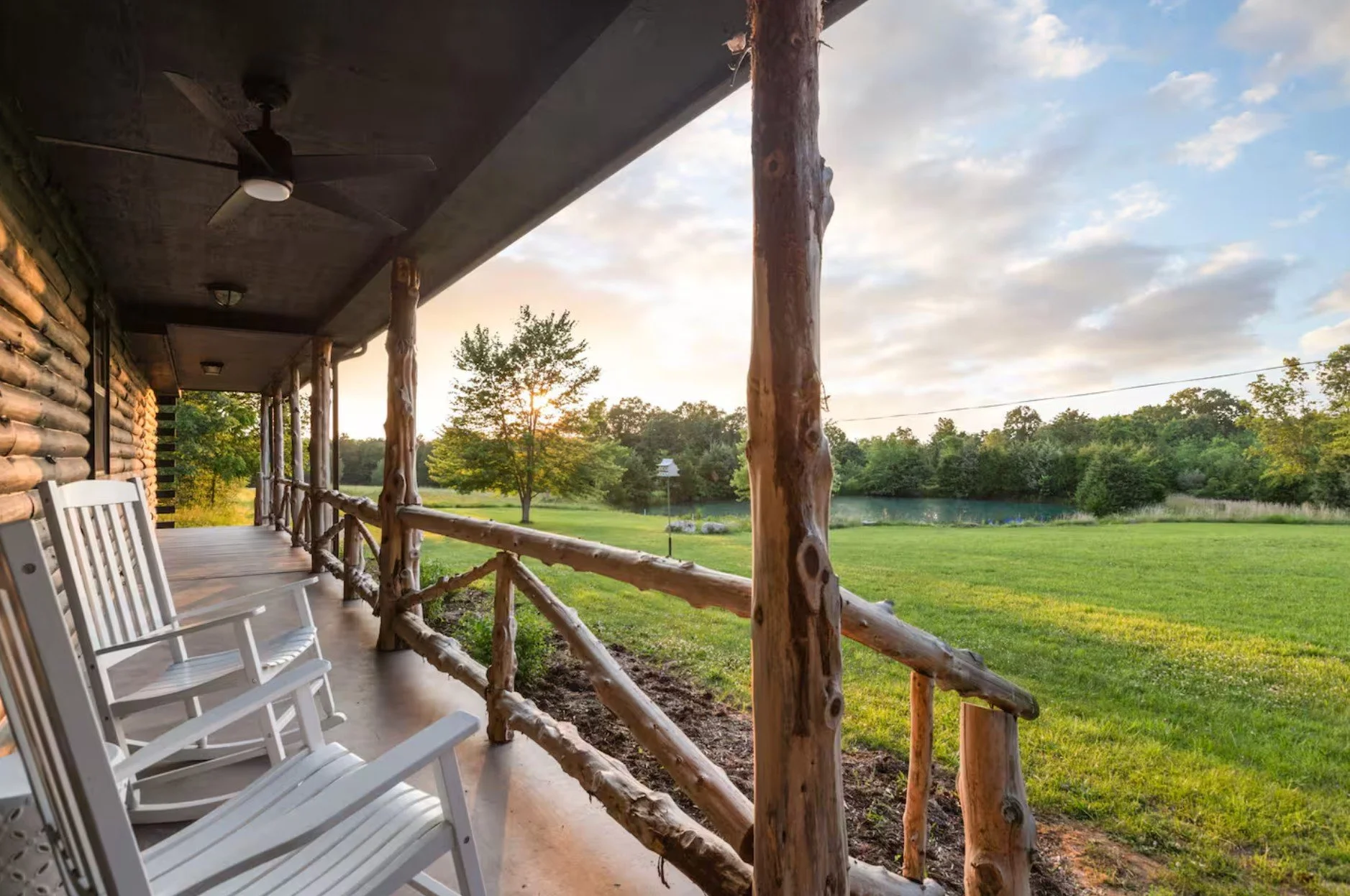 View of a rustic porch with wooden railings and white rocking chairs overlooking a lush green lawn, trees, and a pond at sunset.