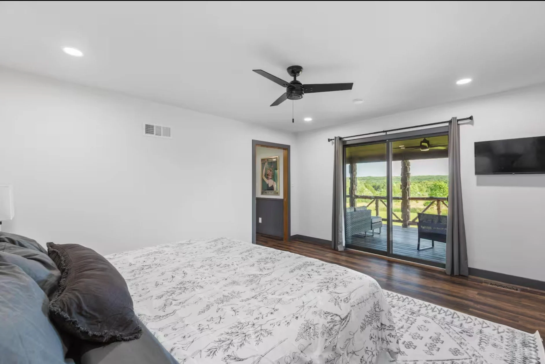 Bedroom with white walls, wooden floor, king-sized bed with white floral bedding, gray and black pillows, ceiling fan, sliding glass door leading to a balcony with outdoor seating, TV mounted on wall, framed artwork, and gray curtains.