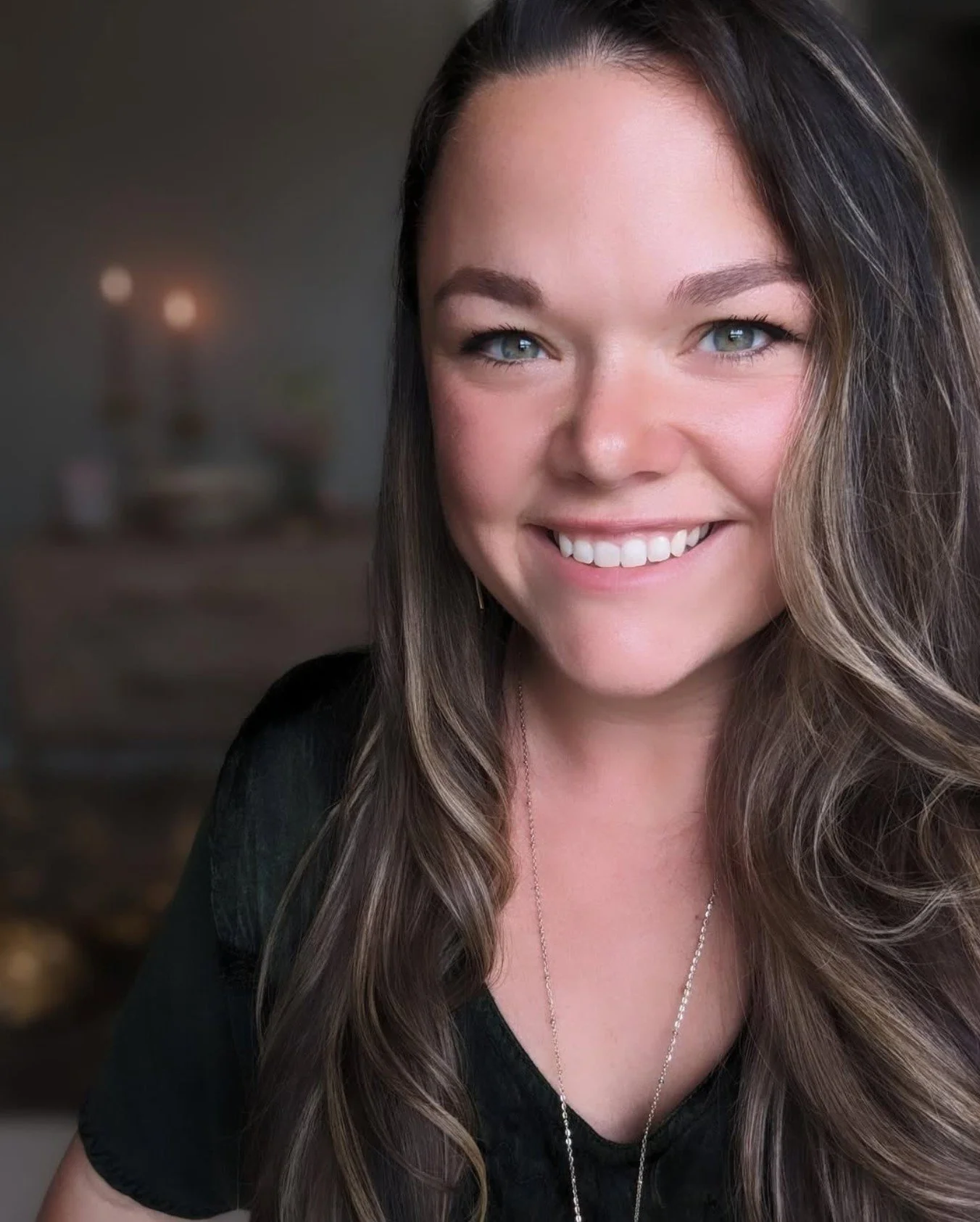 Close-up photo of a smiling woman with long brown hair and blue eyes, wearing a black top and a delicate necklace, with a blurred background.