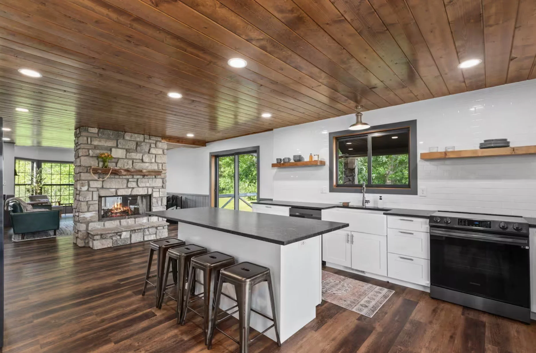 Modern kitchen with white cabinets, black countertops, and a large island with four stools. A stone fireplace is visible in the adjacent living area with a fire burning. Wooden ceiling panels, hardwood floors, and large windows with view of green out