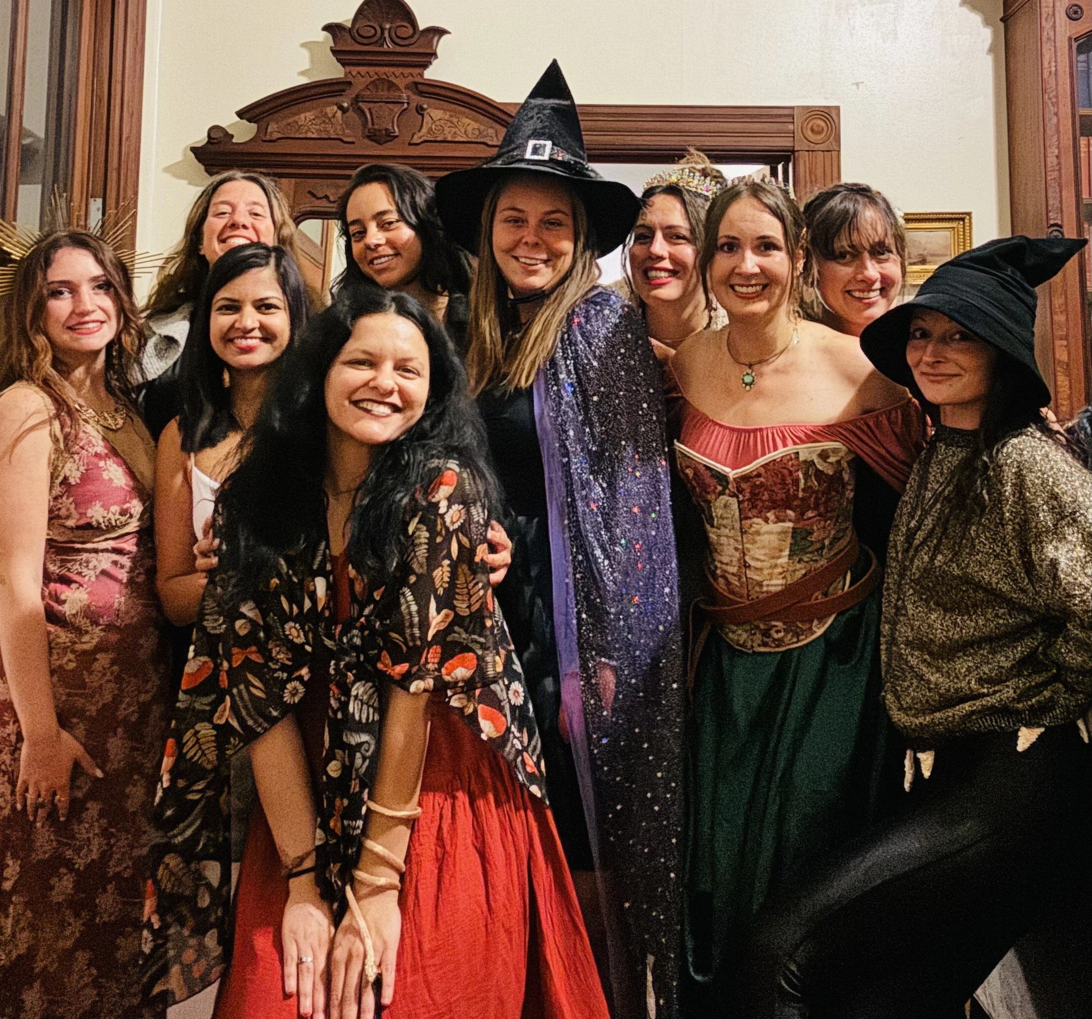 Group of women dressed in Halloween costumes, some wearing witch hats and other fantasy-themed attire, smiling at a party.