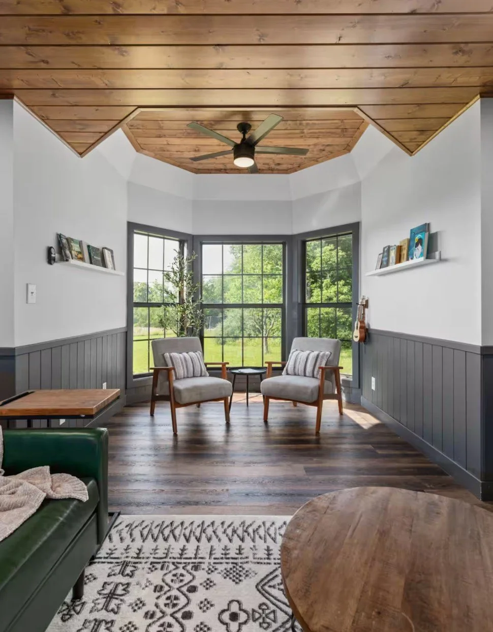 Living room with large bay window, two armchairs with striped pillows, a wooden ceiling fan, and a view of green outdoor scenery.