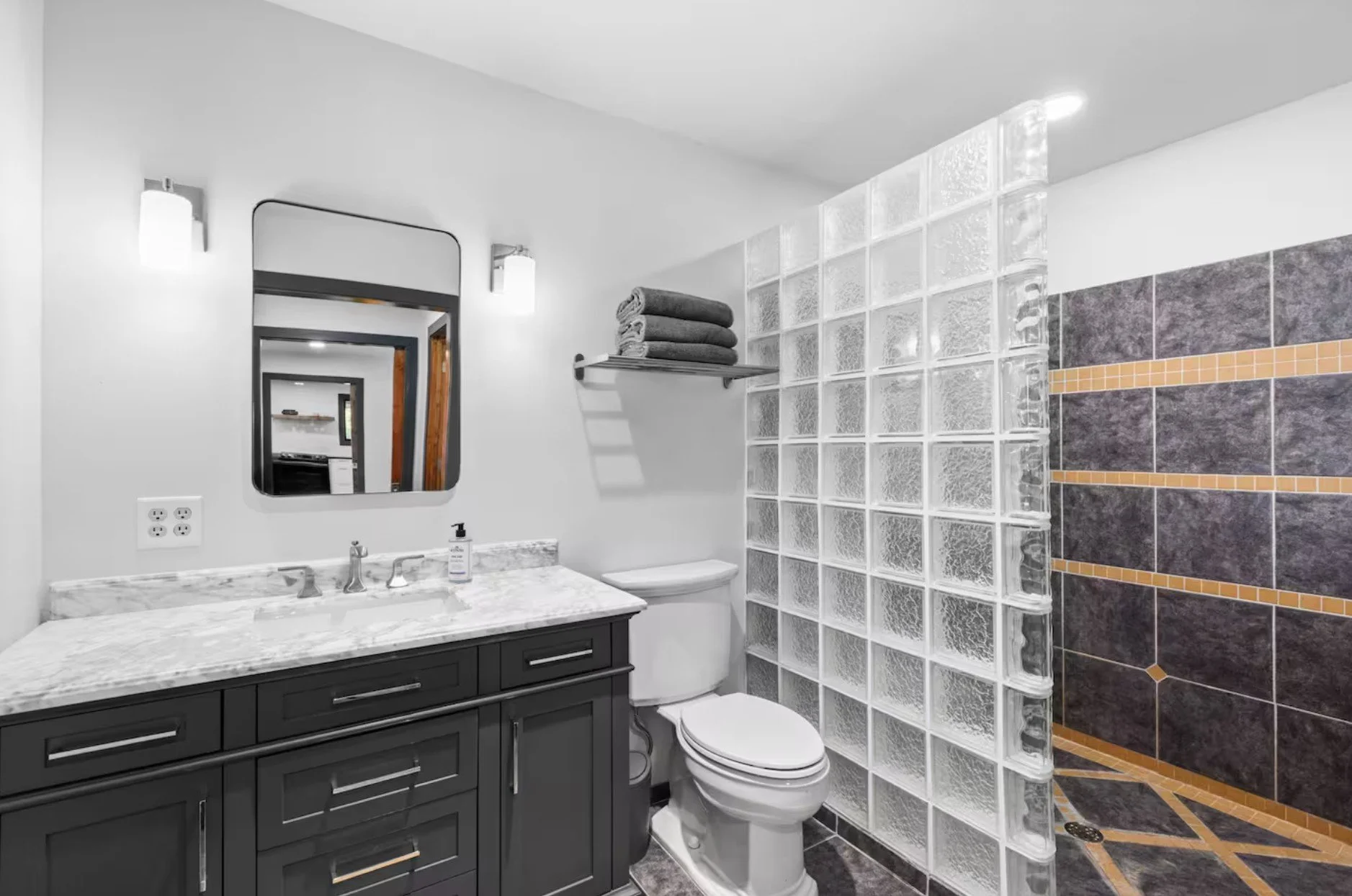 A modern bathroom with a dark gray vanity, marble countertop, and a rectangular mirror. There are two wall-mounted lights, a toilet, and a towel rack with folded towels. The shower area features glass blocks and dark tiled walls with orange accents.