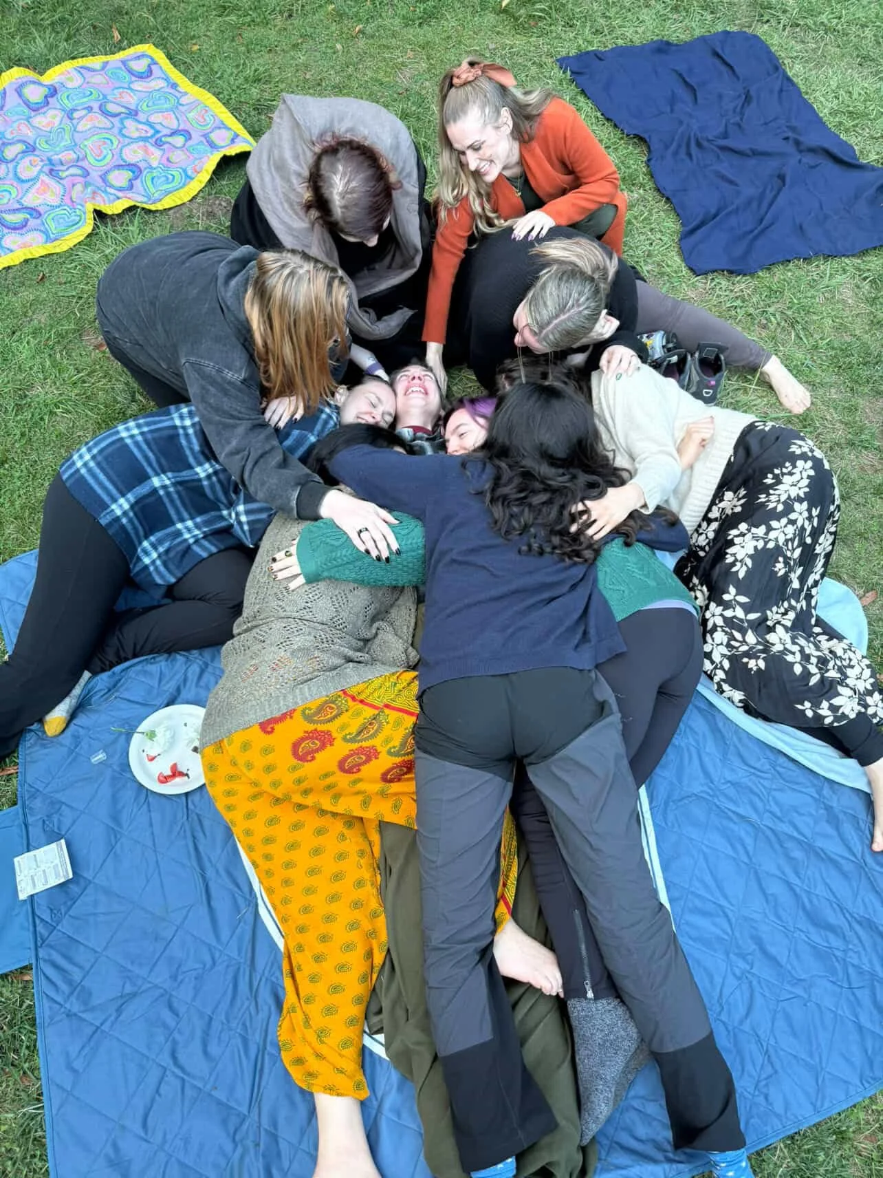 A group of women, are lying and sitting on blankets and mats on the grass, laughing and hugging each other in a lively outdoor gathering.