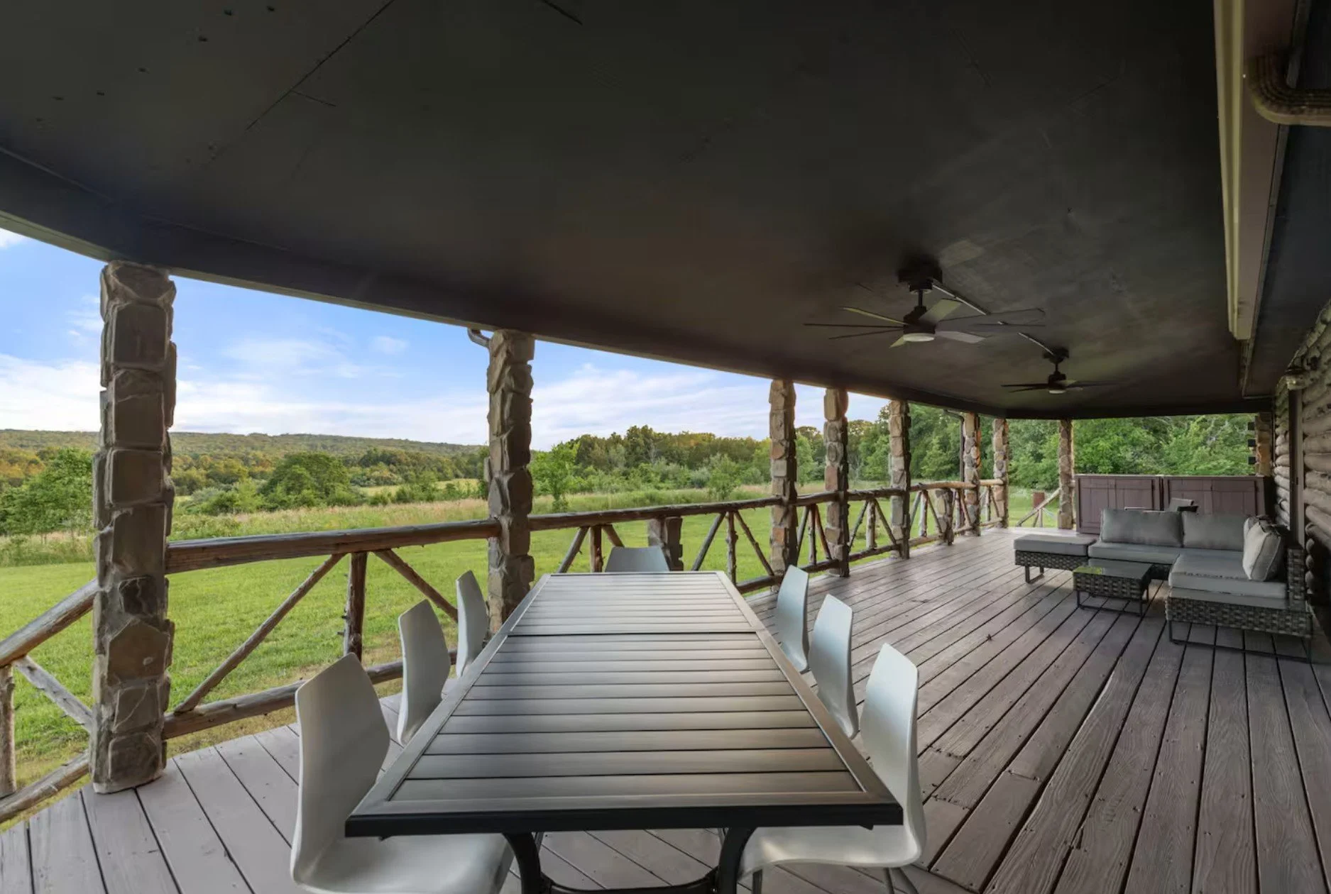 Covered porch with wooden railing, outdoor dining table with six white chairs, cushioned outdoor sofa, ceiling fans, overlooking green landscape with trees and hills.