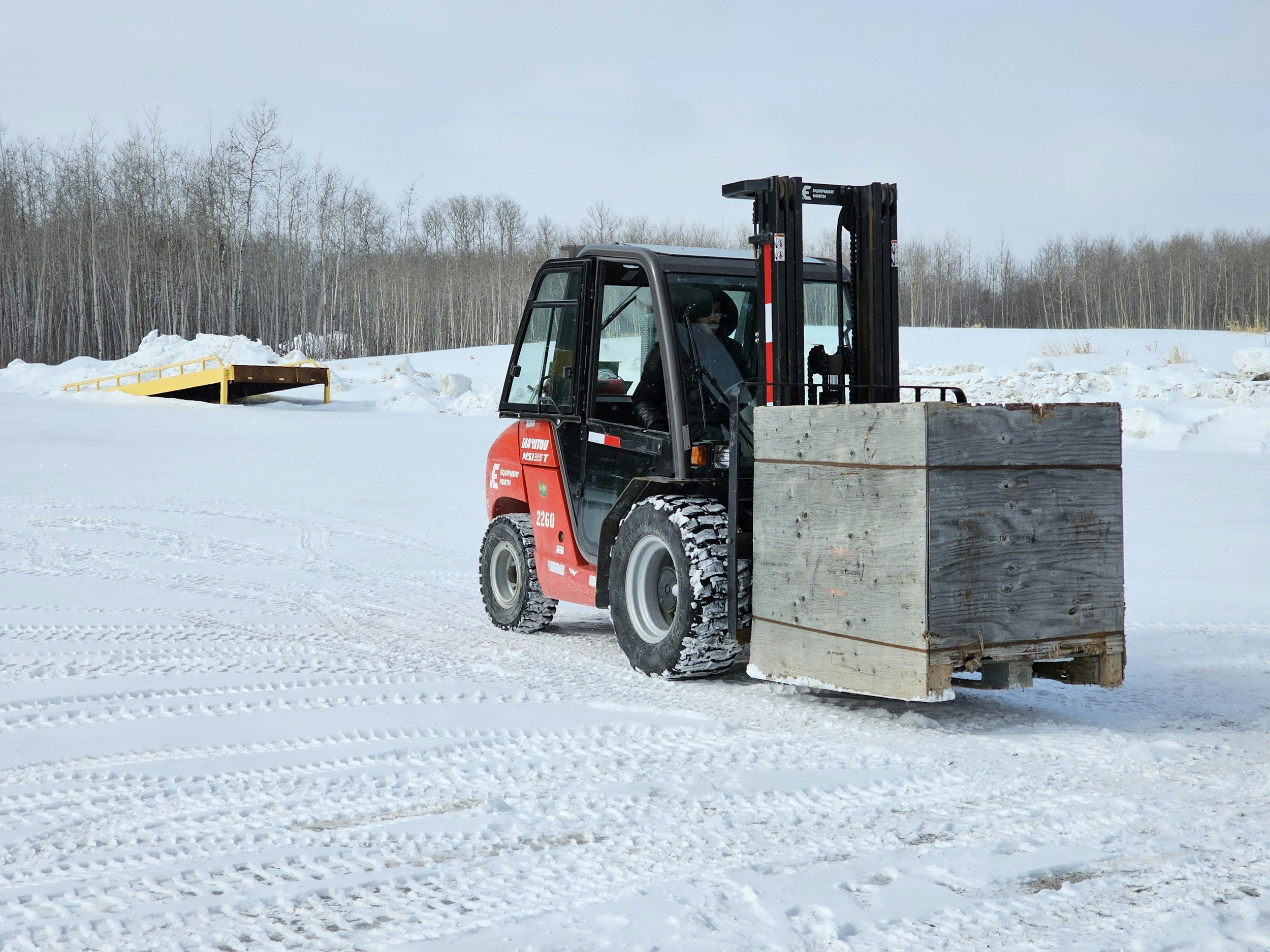 A forklift carrying a large wooden crate in a snowy outdoor area with snow-covered ground and a line of bare trees in the background.