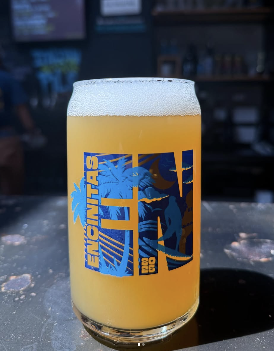 A glass of hazy yellow beer with a foamy head, featuring a blue tropical-themed logo with palm trees, a surfboard, and a person in swimwear, reading "Encinitas" and "2020."