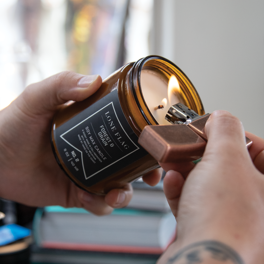 Person lighting a candle from a jar labeled 'Love Foliage' with a lighter, in a room with natural light.