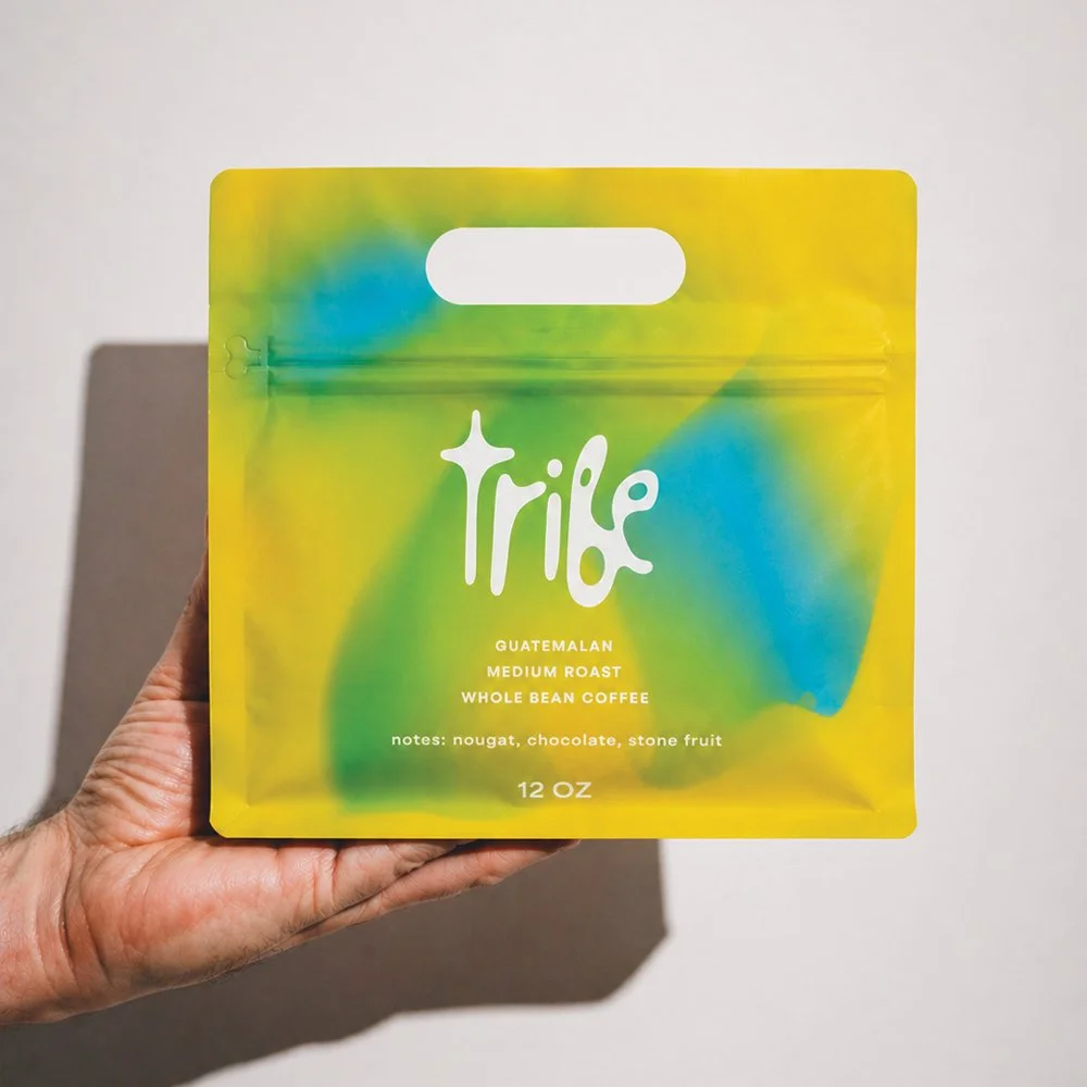 Colorful packaging of Tribe Guatemala medium roast whole bean coffee with notes of nougat, chocolate, and stone fruit, held in a person's hand against a plain background.