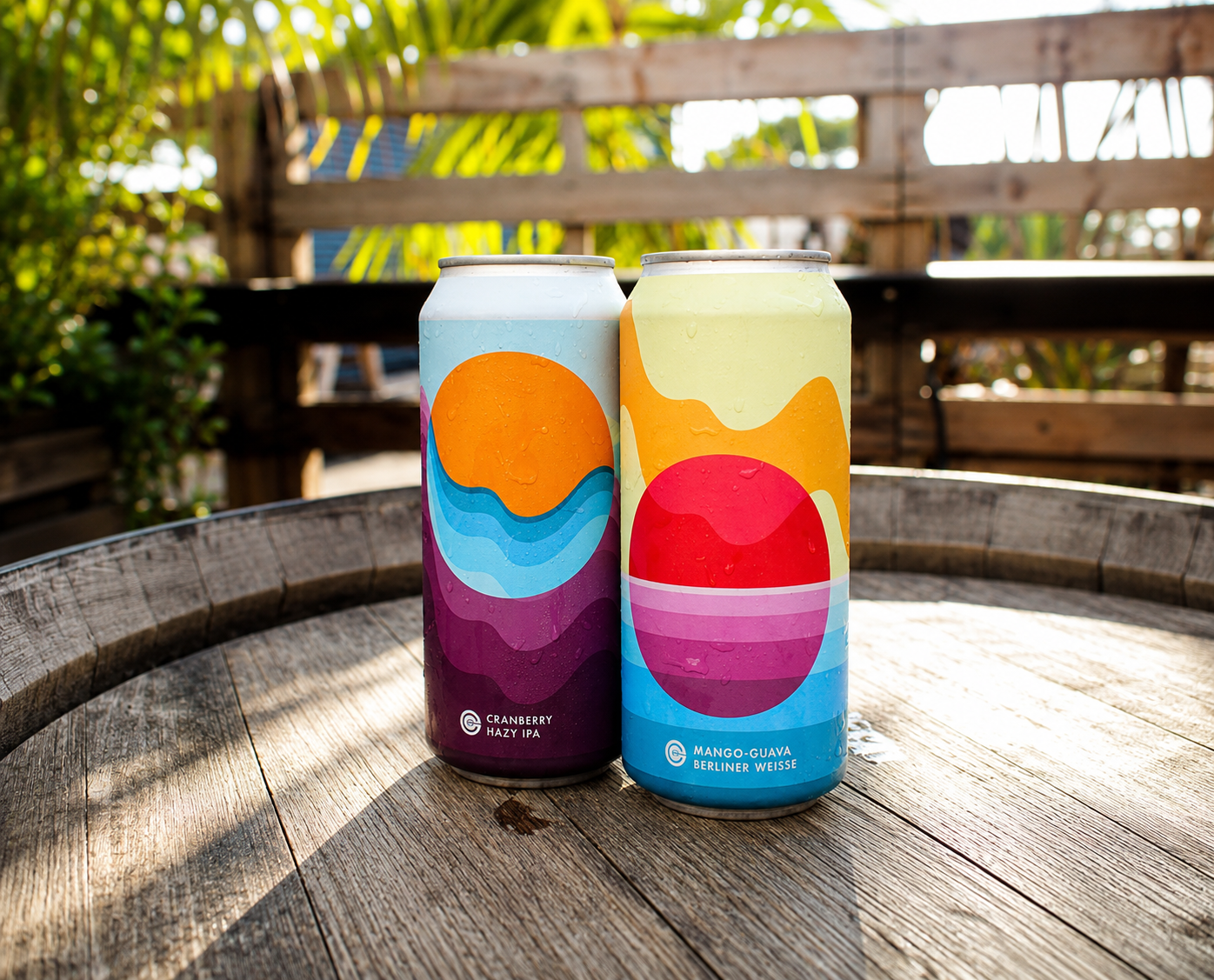 Two colorful cans of beer with abstract art designs sit on a wooden table outdoors, with a wooden fence and green foliage in the background.