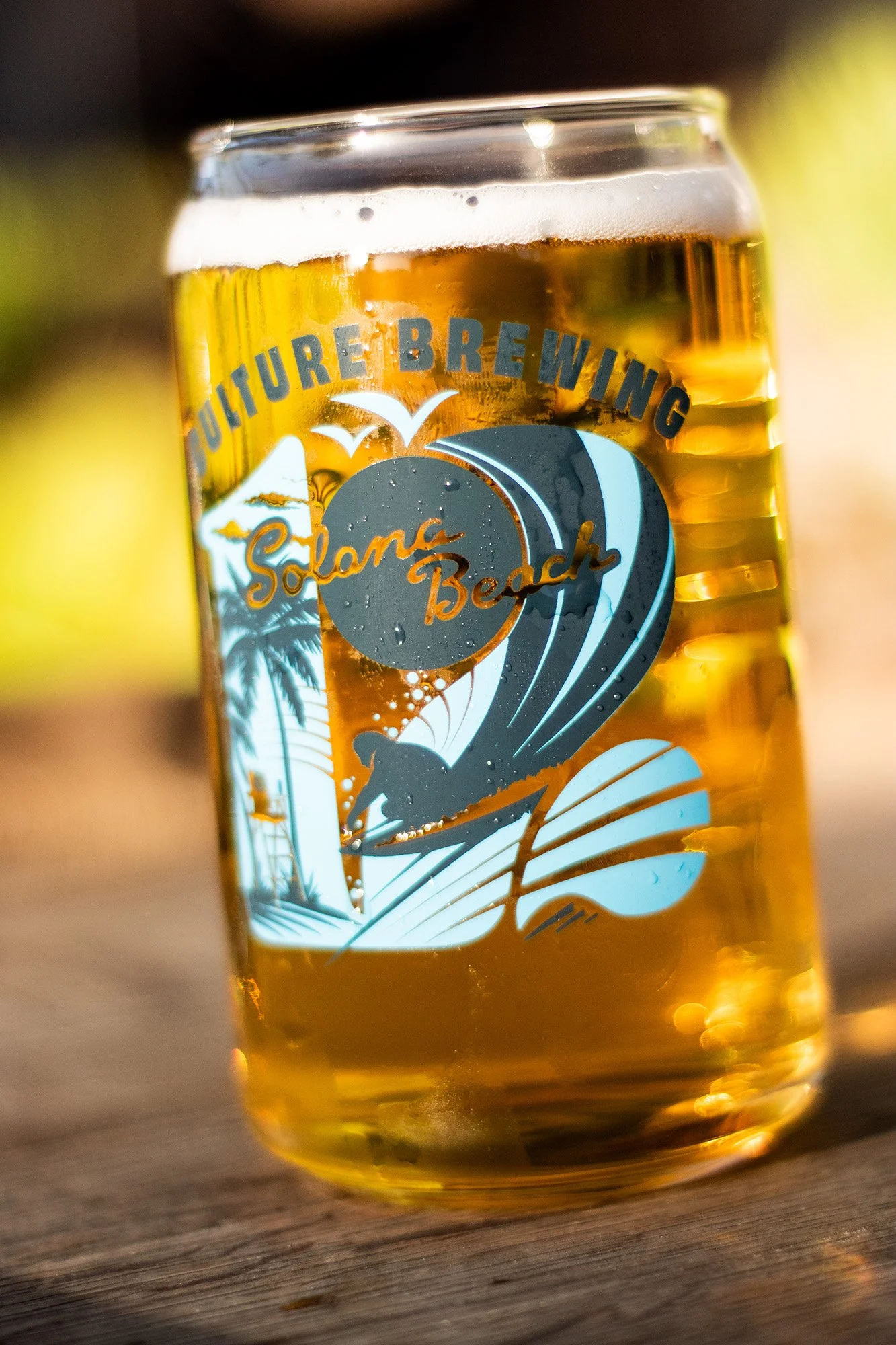 A clear glass filled with golden beer, displaying a logo of a surfing scene with a palm tree and waves, with the words 'Suriture Brewing' and 'Shores Beach'.