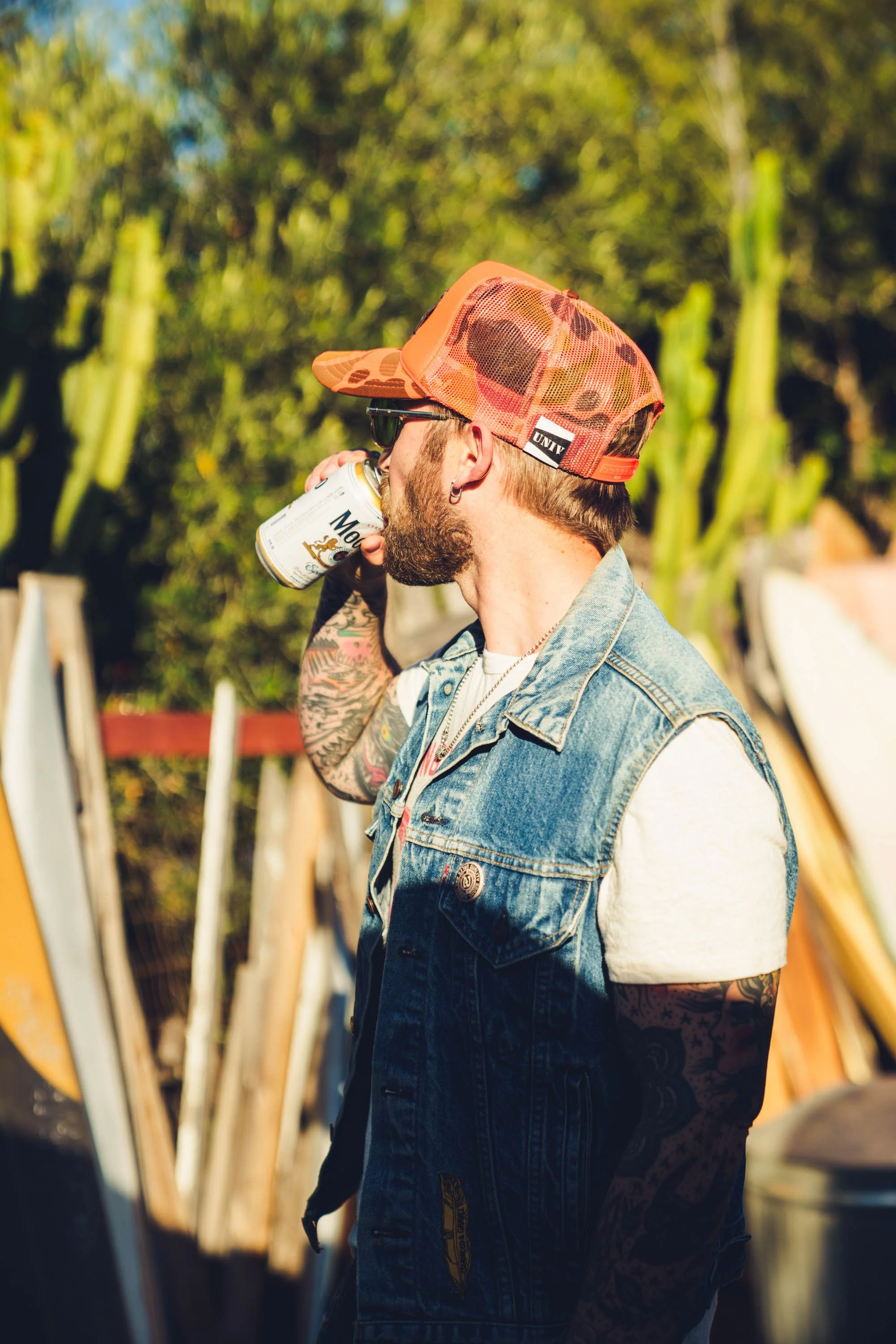 A man with tattoos, a beard, and earrings wearing a floral cap, sunglasses, and a denim vest drinking from a can in an outdoor setting with plants and trees in the background.