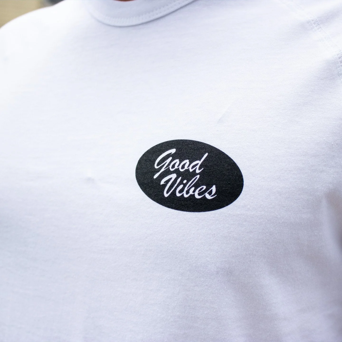 Close-up of a white T-shirt with a black oval graphic containing the words "Good Vibes" in white cursive font.