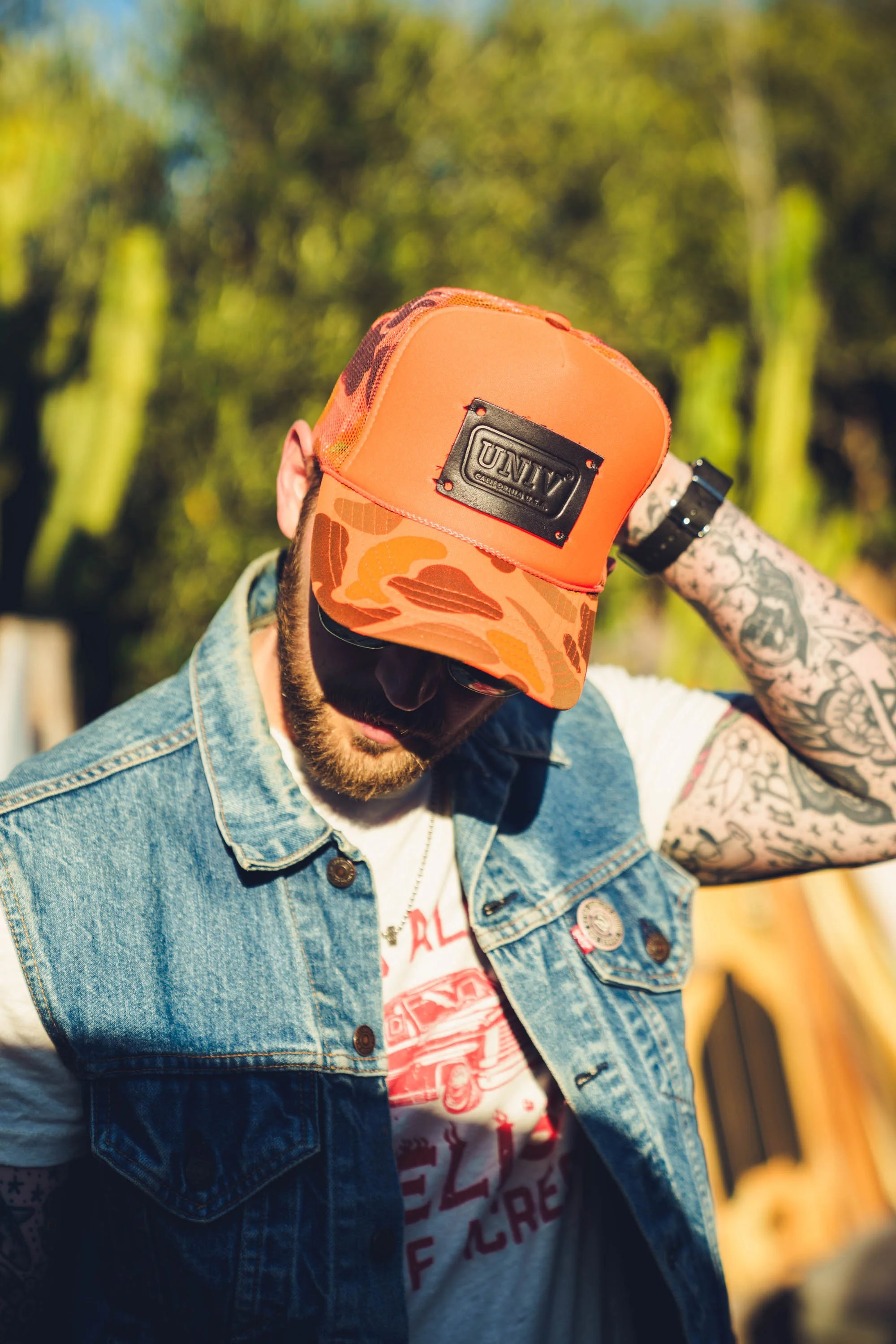 A man with tattoos wearing a denim vest and a red trucker hat with a black patch, adjusting his hat with his right hand. The background is outdoors with green foliage and yellow structures.