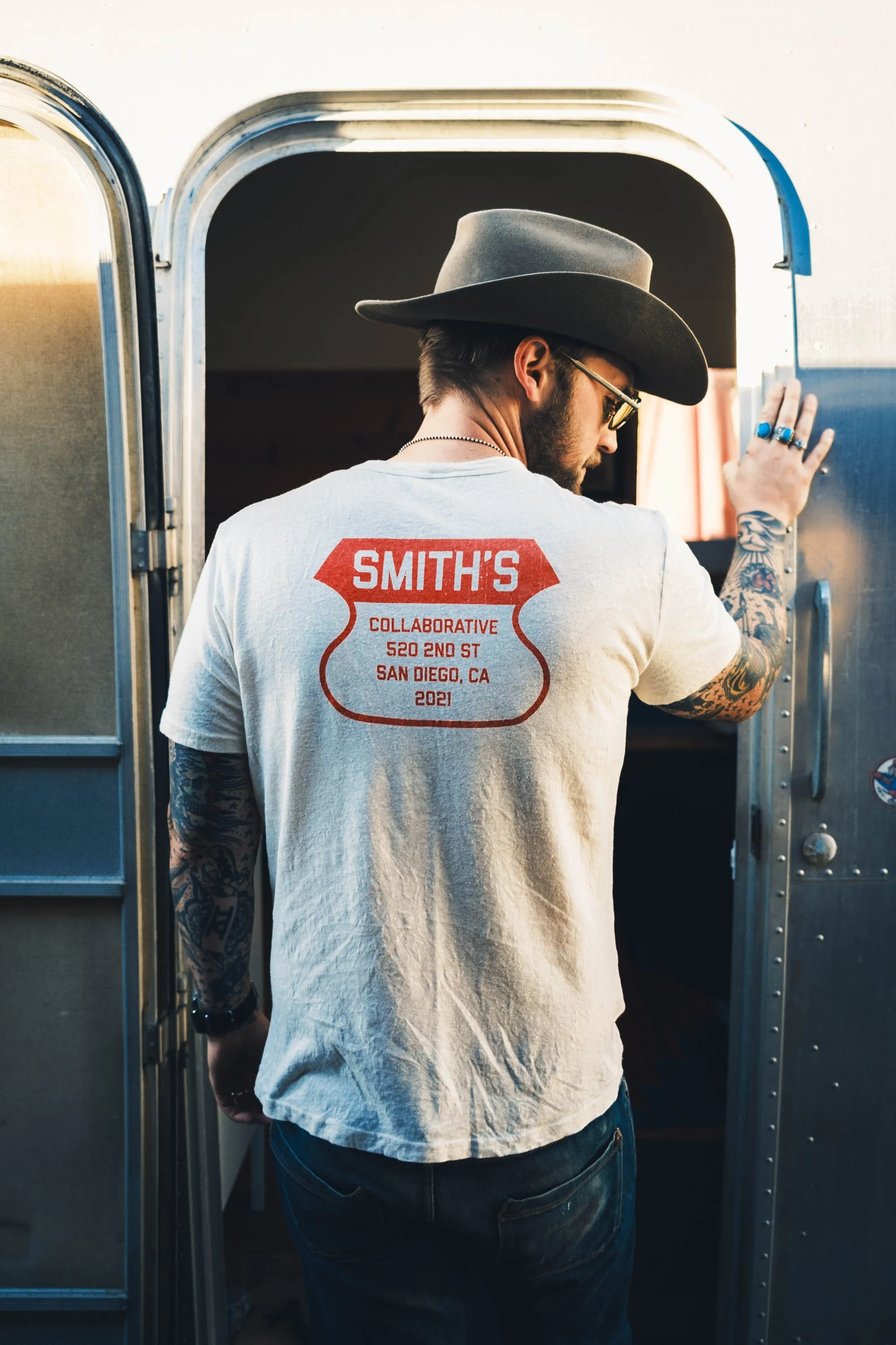 A man wearing a wide-brimmed hat, sunglasses, and a white t-shirt with red text, standing at an open doorway of a silver trailer, with tattoos on his arms.