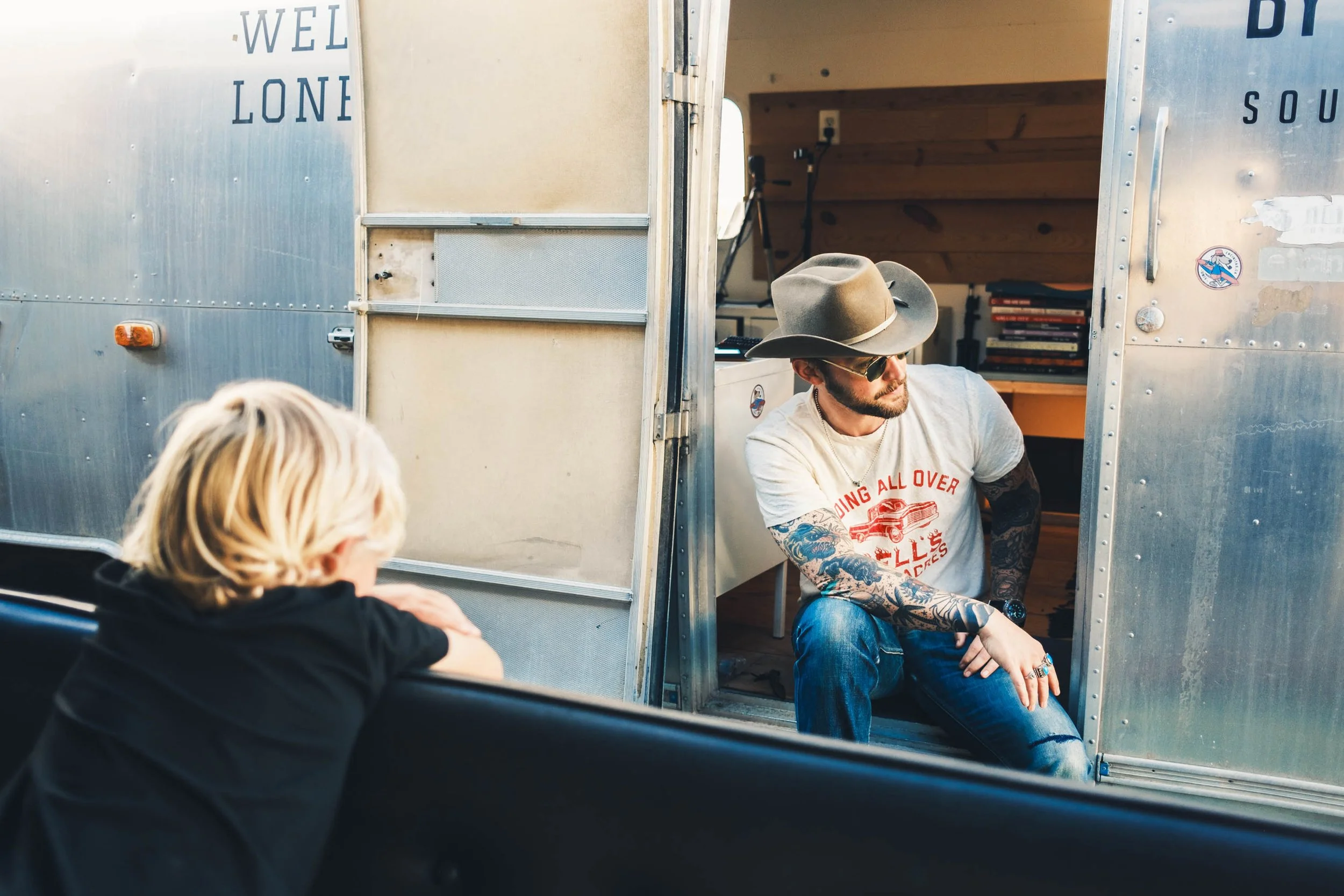 Man wearing a wide-brimmed hat, sunglasses, and a graphic t-shirt with tattoos on his arms, sitting outside a vintage trailer with a wooden interior, while a person with blonde hair is visible in the foreground.