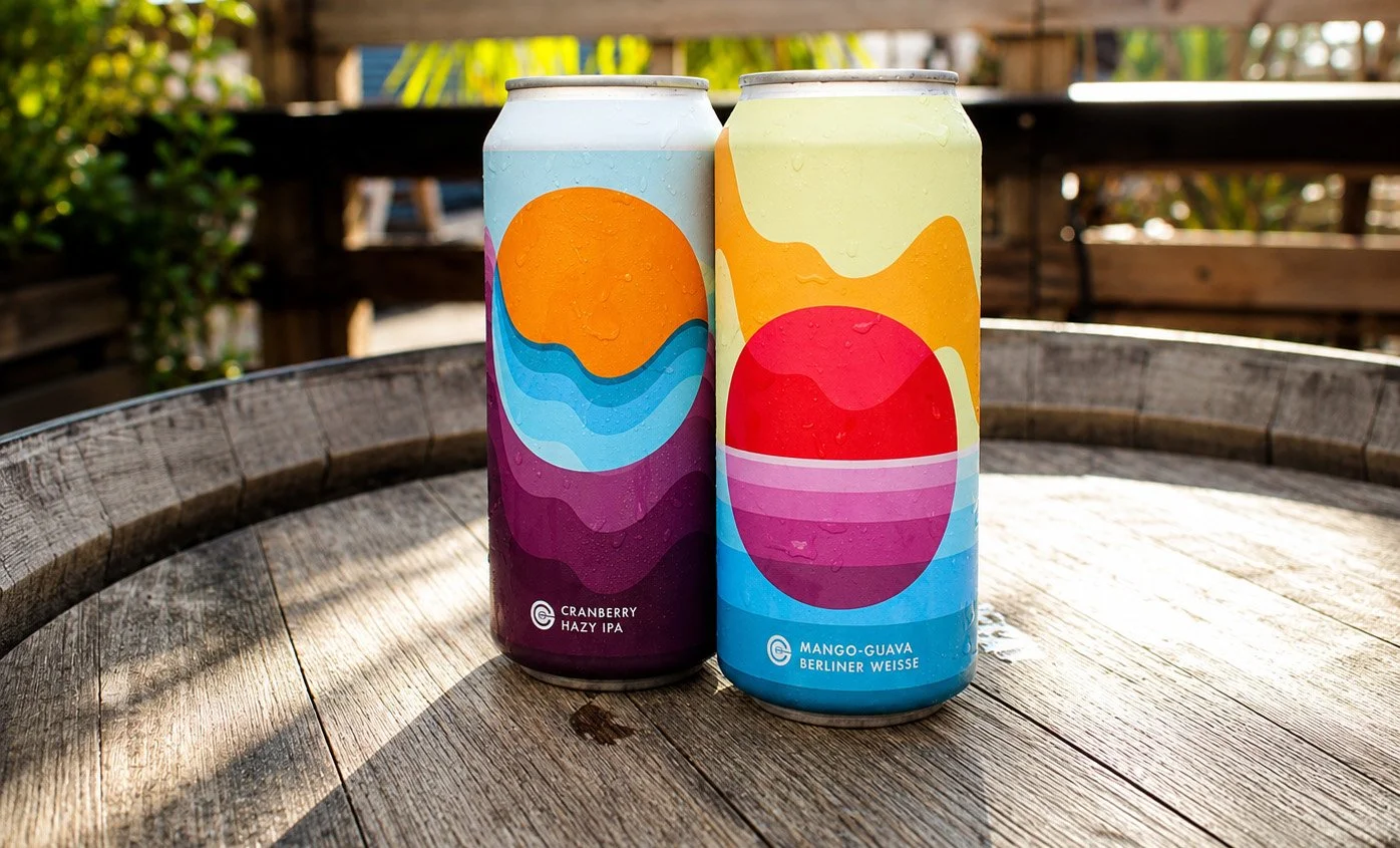 Two colorful cans of craft beer on a wooden outdoor table with a blurred background of greenery and wooden railing.