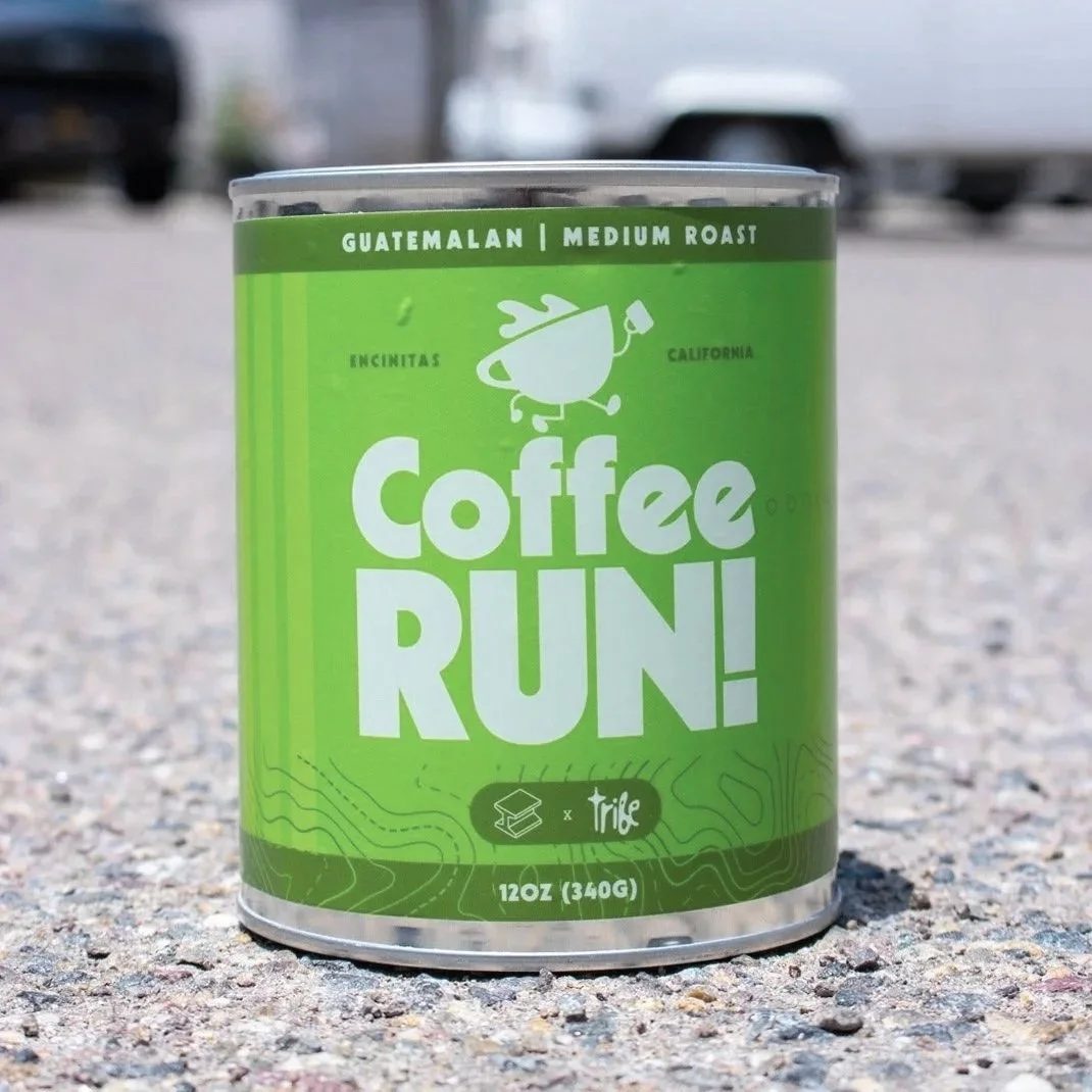 A can of Guatemala medium roast coffee labeled "Coffee Run!" placed on gravel pavement with cars and a parking lot in the background.