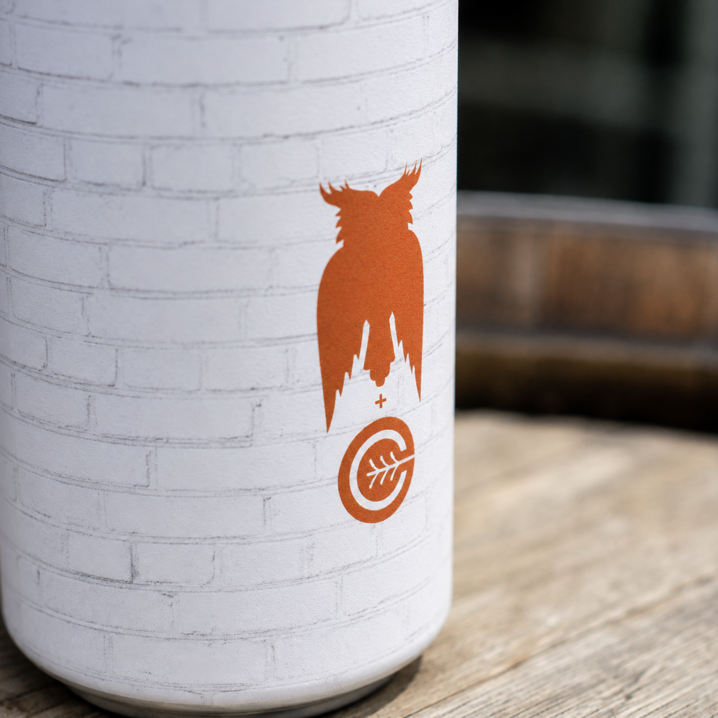 Close-up of a white brick wall with an orange graphic of an owl with outstretched wings, a plus sign, and a target symbol with a leaf inside, on a cylindrical surface.