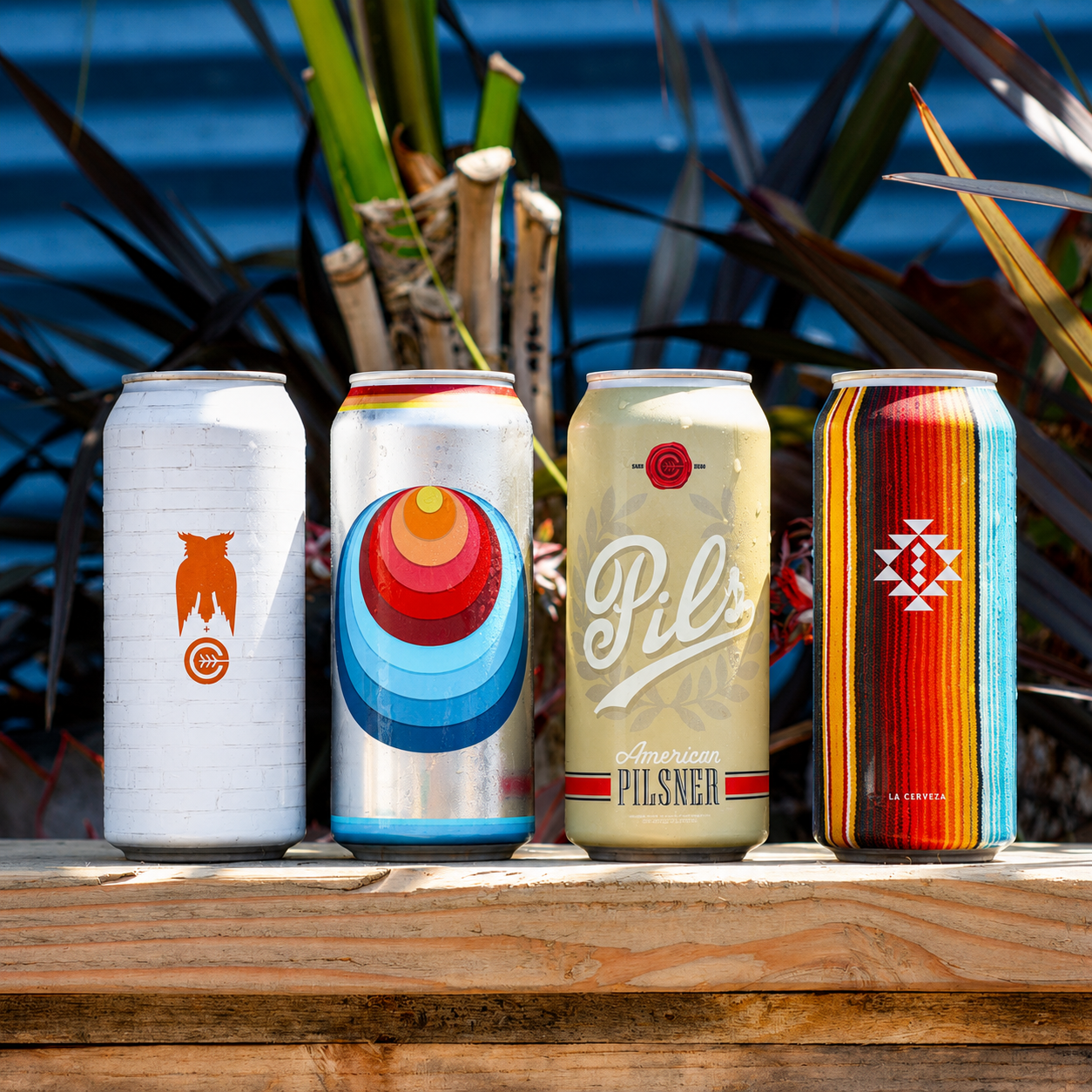 Four cans of beer with colorful designs on a wooden surface outdoors, background of green plants and water.
